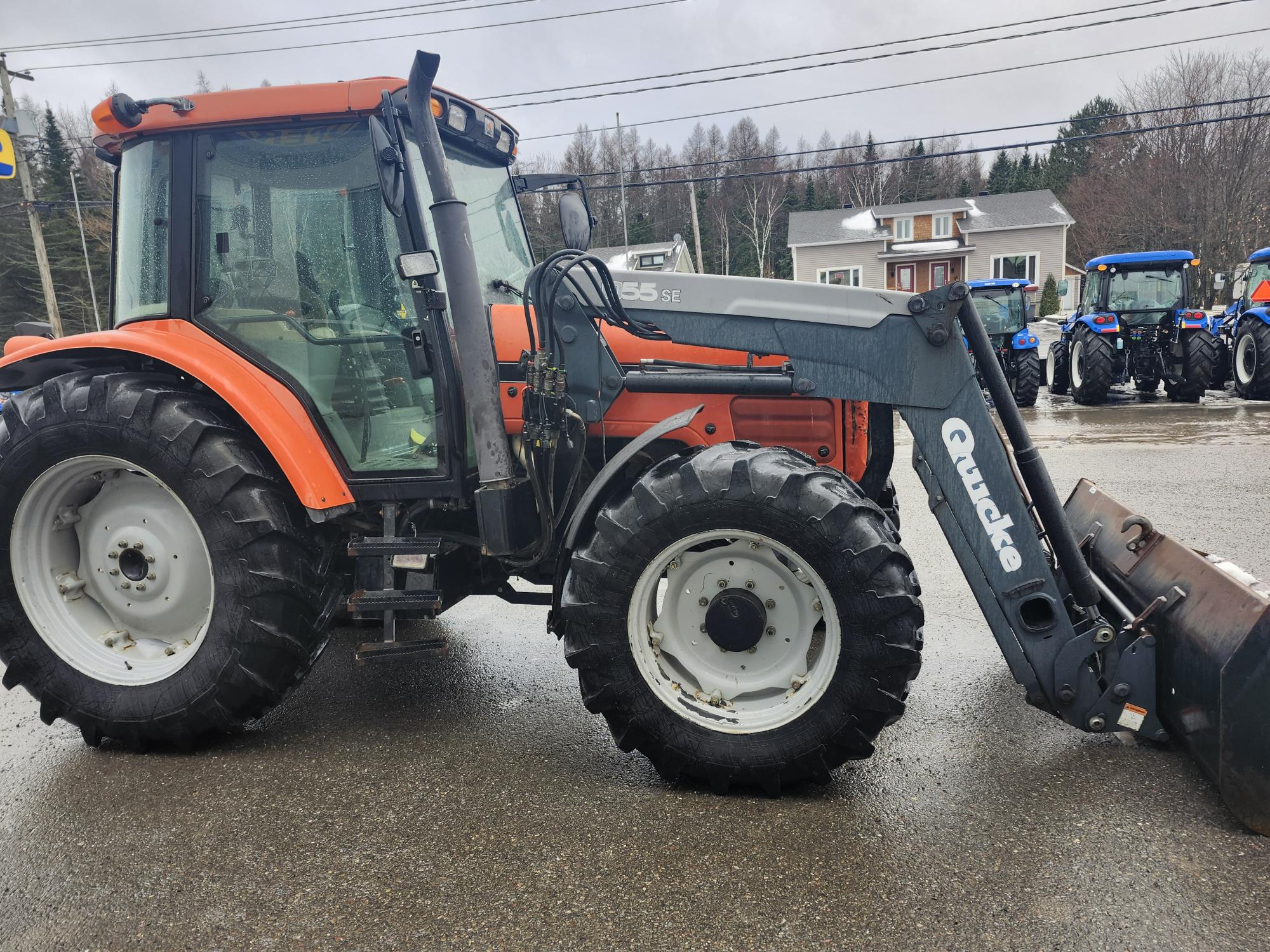 Un tracteur rouge équipé d'un chargeur frontal est stationné sur une route mouillée. Plusieurs autres tracteurs sont visibles en arrière-plan près d'une zone résidentielle avec des arbres et un ciel couvert.