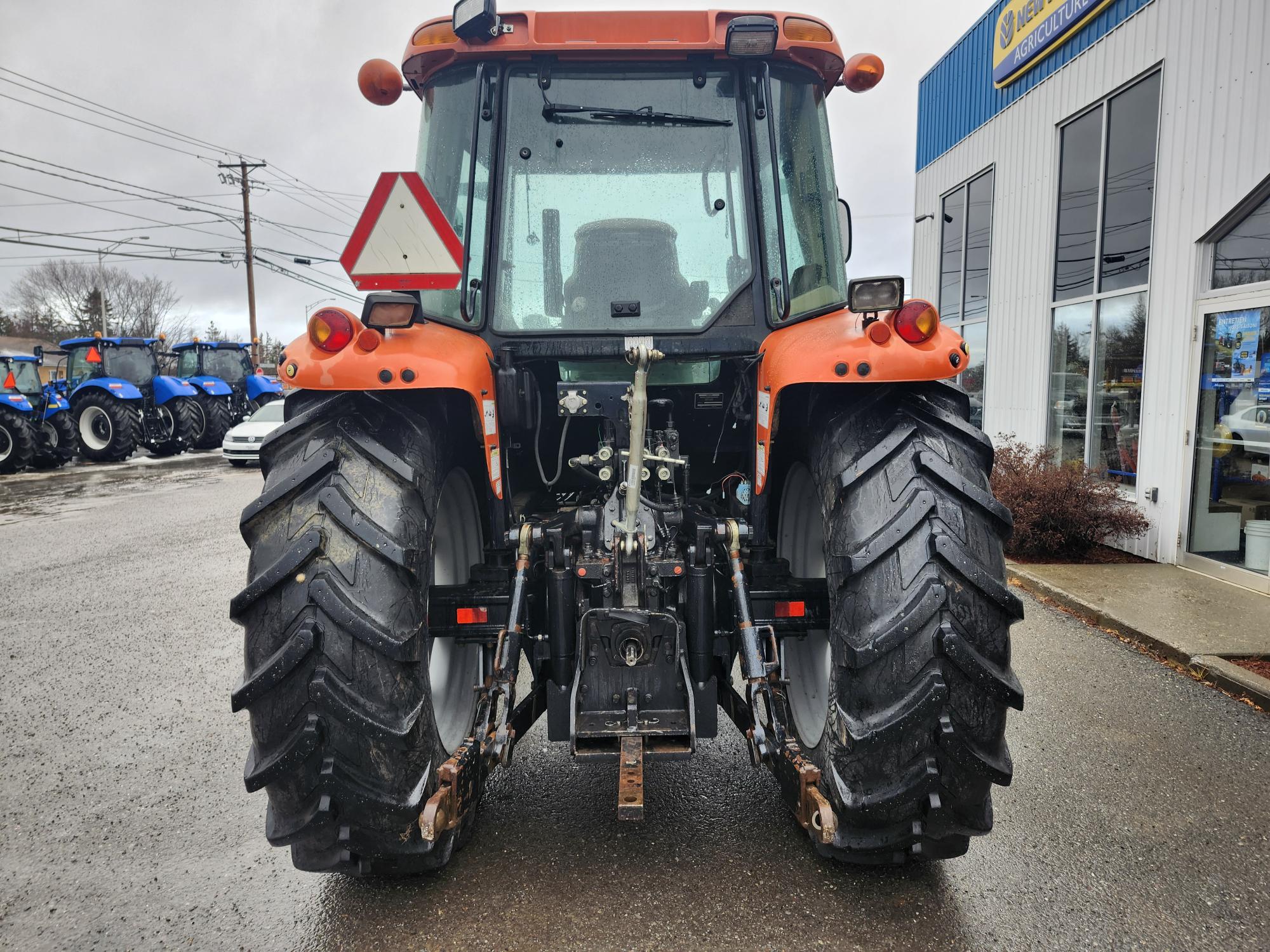 Vue arrière d'un tracteur orange stationné sur un trottoir mouillé à l'extérieur d'un concessionnaire. Le tracteur est équipé de gros pneus noirs et de feux arrière visibles, avec un panneau réfléchissant triangulaire monté à l'arrière.