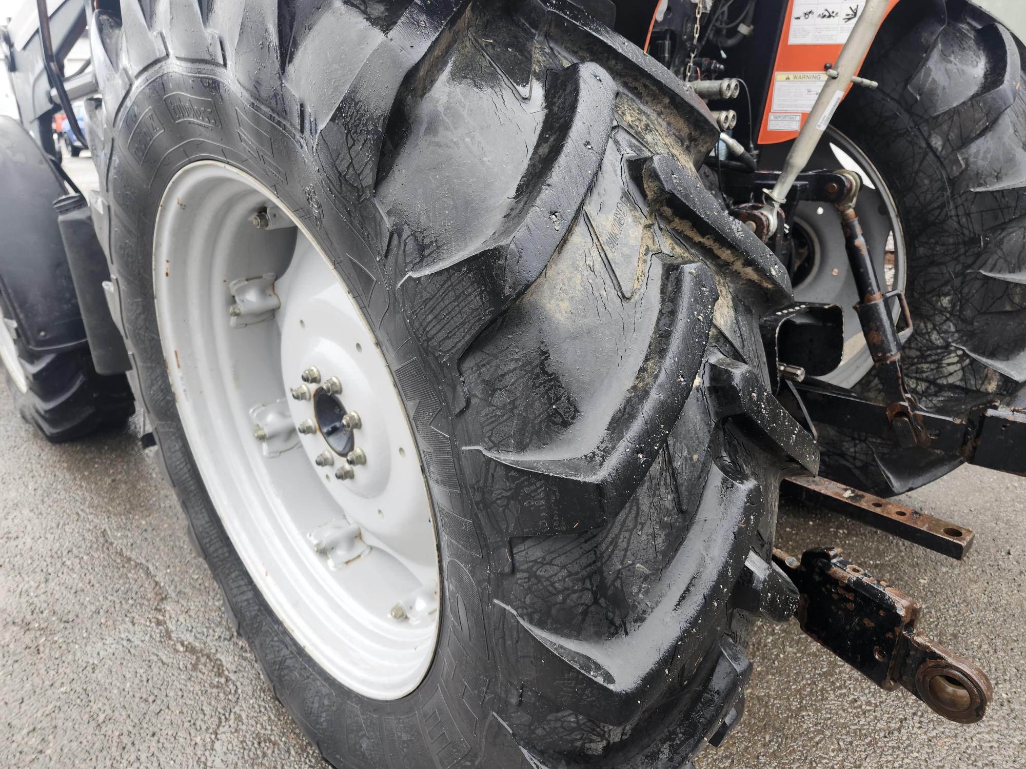 Gros plan d'un gros pneu arrière de tracteur sur une surface mouillée. La bande de roulement est visible, mettant en valeur la texture rugueuse et usée. Le bord du tracteur et une partie de sa machinerie sont également visibles à l'arrière-plan.