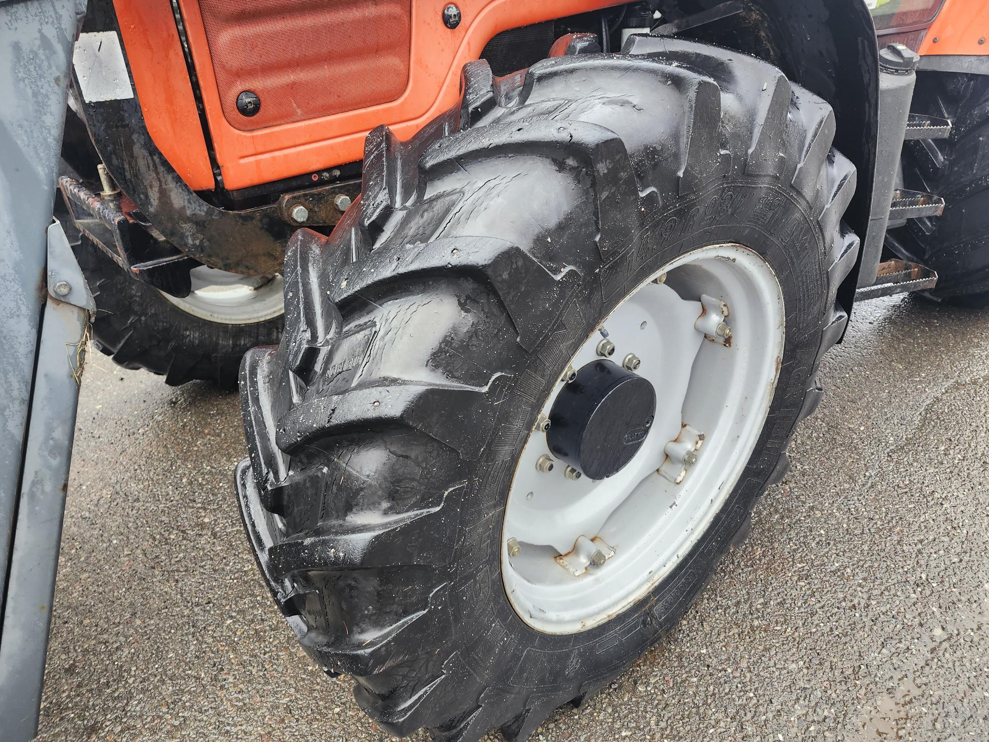 Gros plan d'un pneu arrière de tracteur aux sculptures profondes, posé sur une surface pavée et mouillée. La carrosserie orange du tracteur est partiellement visible à l'arrière-plan.