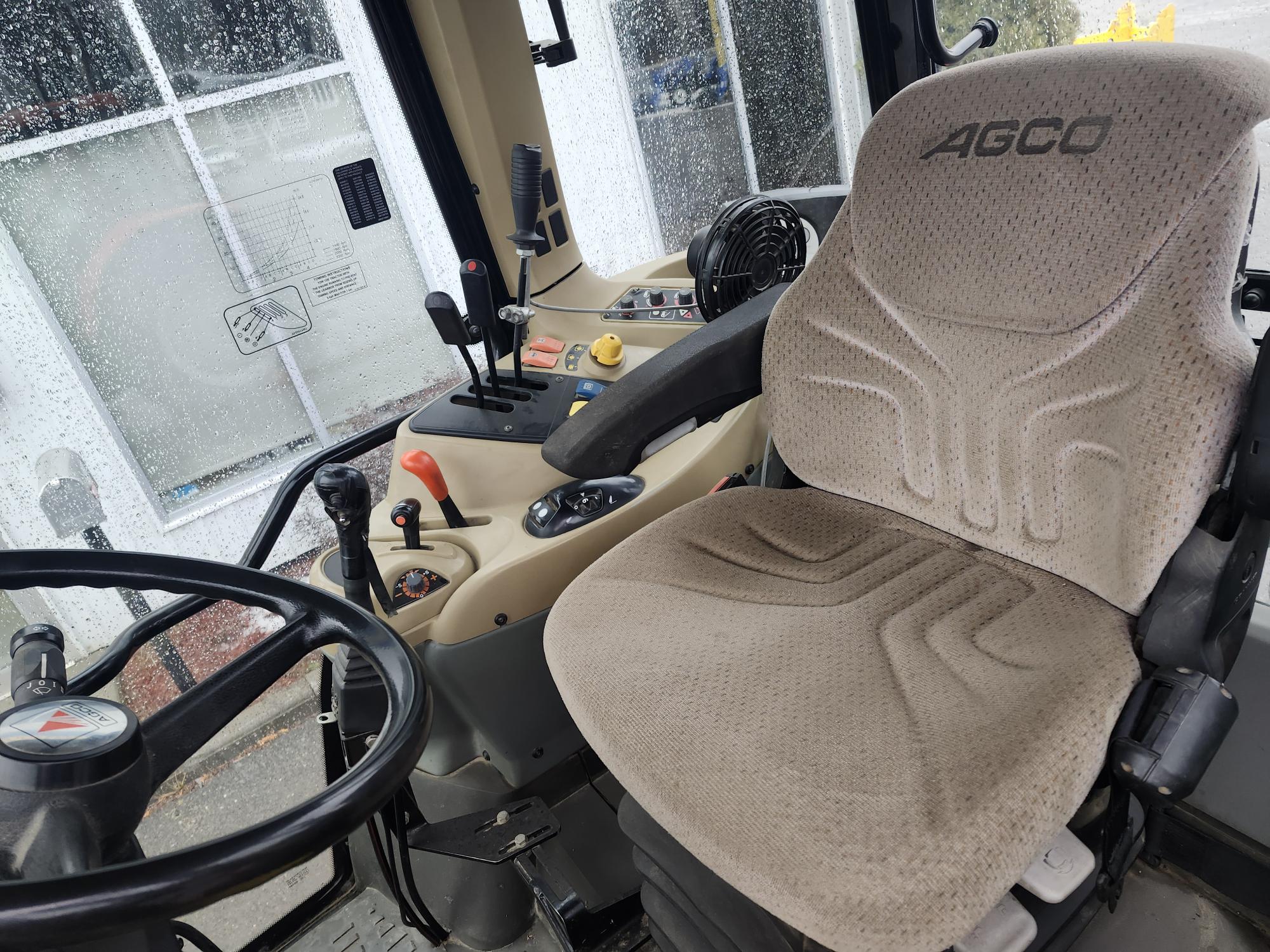 L'image montre l'intérieur d'une cabine de tracteur, comprenant un siège conducteur beige avec la marque « AGCO », un volant, divers leviers et panneaux de commande et une fenêtre avec des gouttes de pluie visibles à l'extérieur.