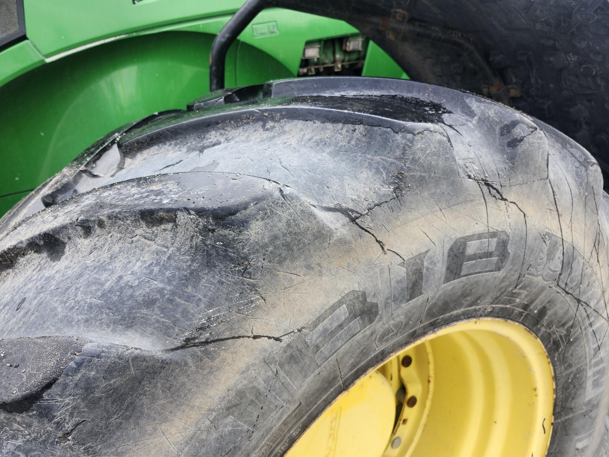 Gros plan d'un gros pneu de tracteur présentant des fissures visibles et une bande de roulement usée, monté sur une roue jaune. Une partie de la carrosserie verte du tracteur est visible en arrière-plan.