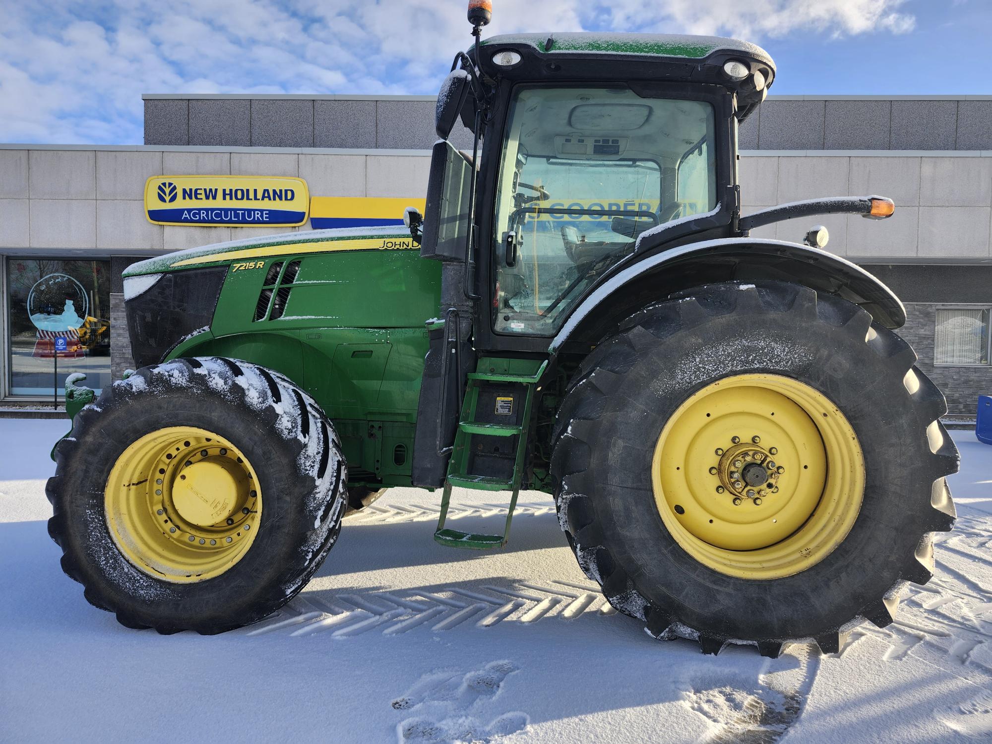 Un tracteur vert et jaune aux pneus recouverts de neige est garé devant un bâtiment de New Holland Agriculture. Le sol est recouvert d'une légère couche de neige et le ciel est partiellement nuageux.