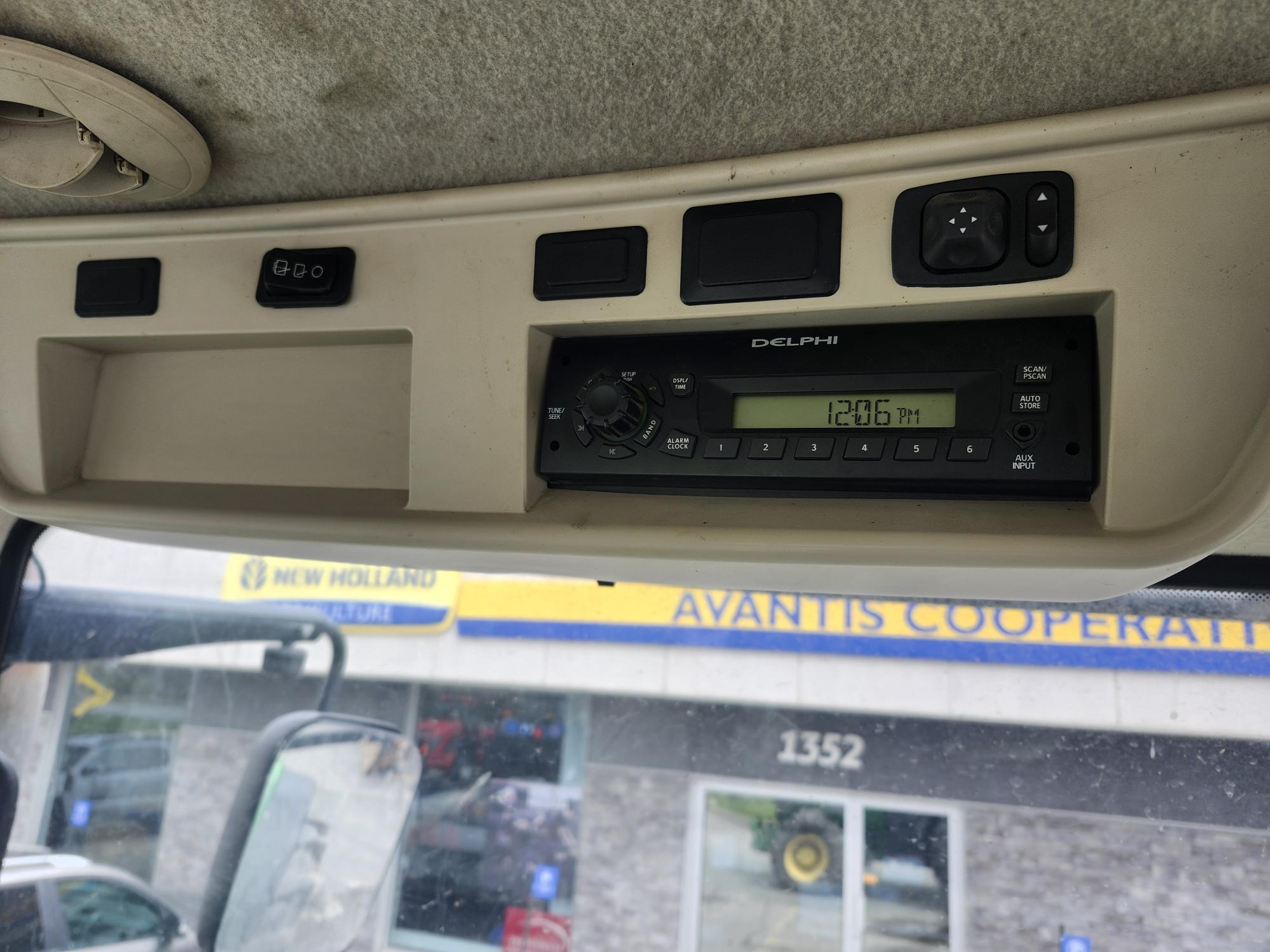 L'image montre la console de pavillon à l'intérieur de la cabine d'un véhicule, avec un écran radio Delphi indiquant 12h36. À l'extérieur, on peut voir par la fenêtre une enseigne de concessionnaire New Holland et un tracteur.