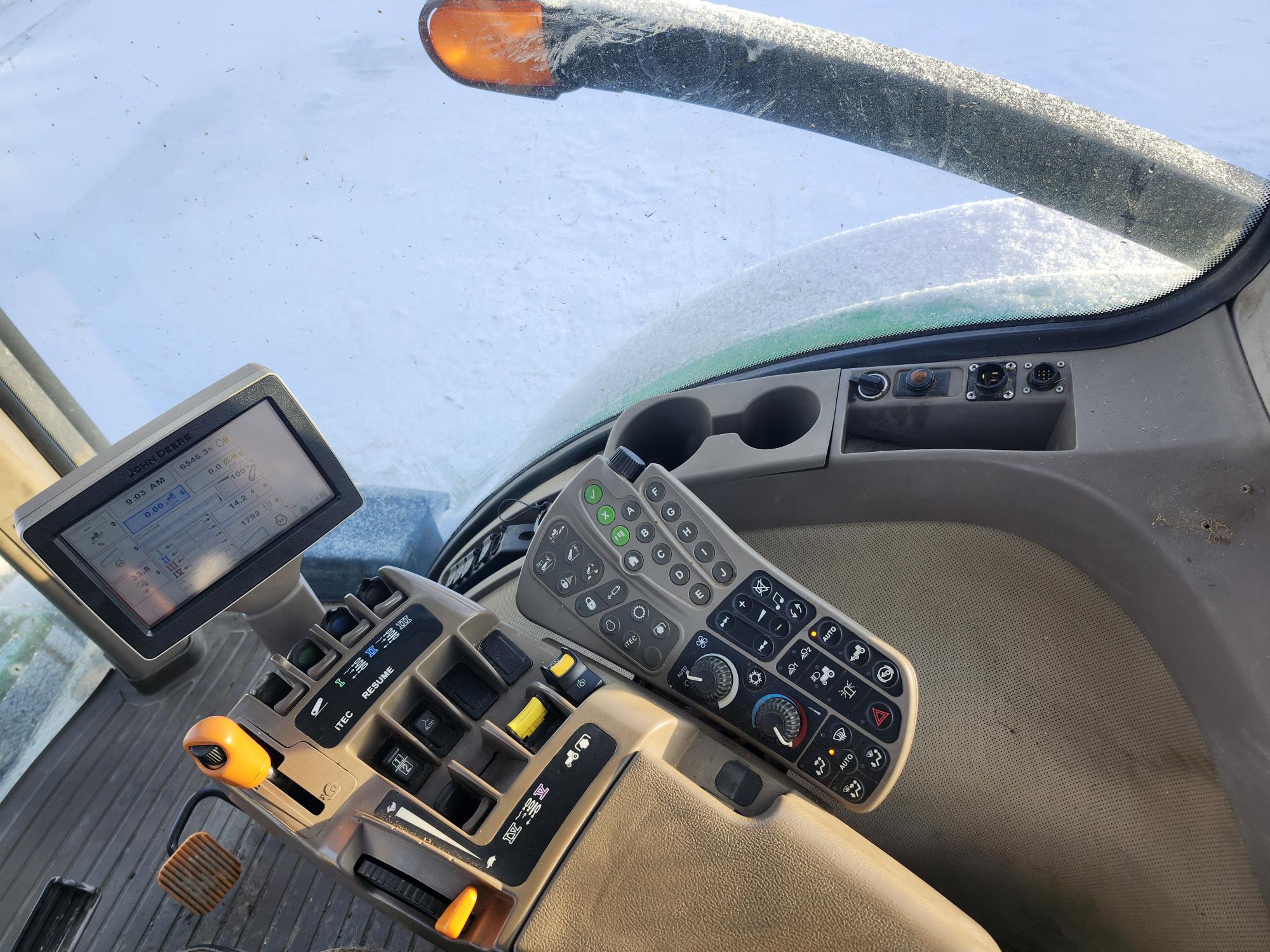 Vue intérieure d'une cabine de tracteur, avec un panneau de commande avec divers interrupteurs, leviers et écran tactile. Un siège est partiellement visible et le pare-brise montre un paysage extérieur enneigé.