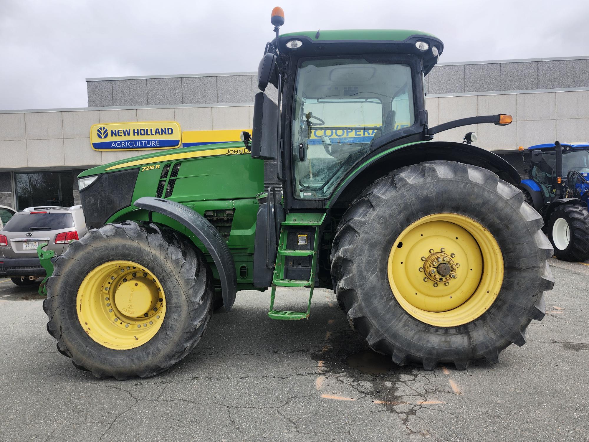 Un tracteur John Deere vert avec de grandes roues jaunes est garé devant un bâtiment portant une enseigne New Holland Agriculture. Le ciel est couvert.