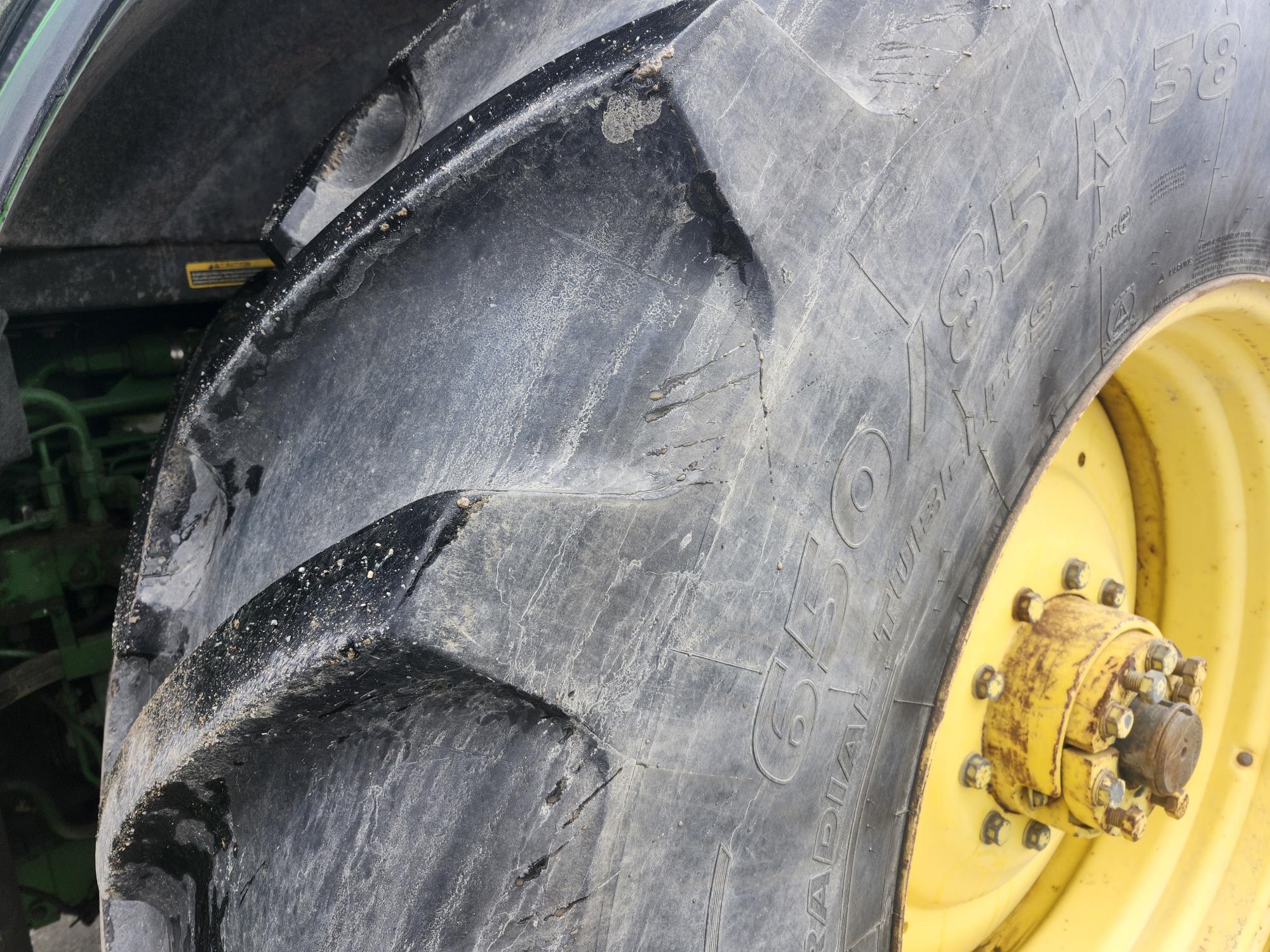 Gros plan d'un gros pneu de tracteur noir usé, aux sculptures profondes, monté sur une roue jaune. Le flanc du pneu présente des lettres et des chiffres visibles. Une partie de la carrosserie verte du tracteur est visible en arrière-plan.