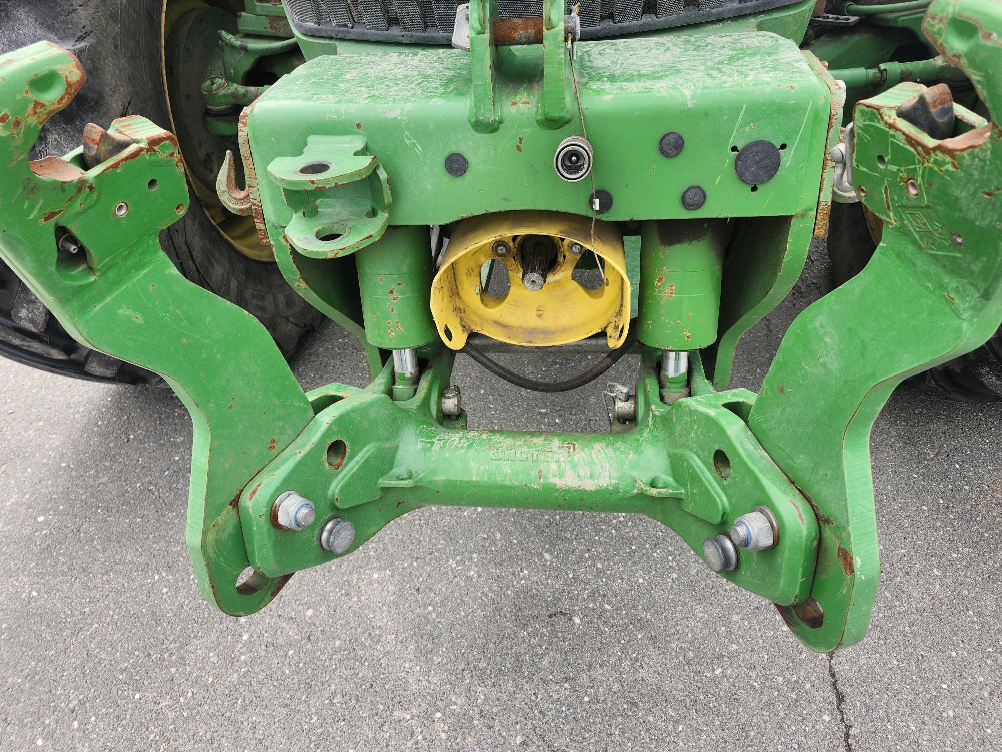Gros plan de l'attelage arrière et de la liaison d'un tracteur agricole vert, montrant des composants métalliques avec de la rouille et des boulons visibles, fixés sur une surface pavée.