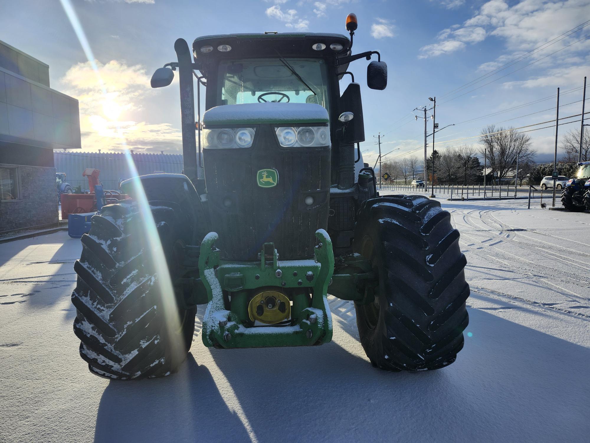 Un tracteur avec de gros pneus est garé sur une surface enneigée. Le soleil brille à l'arrière-plan, projetant une lumière vive. Des lignes électriques et des arbres sont visibles sur le côté droit.