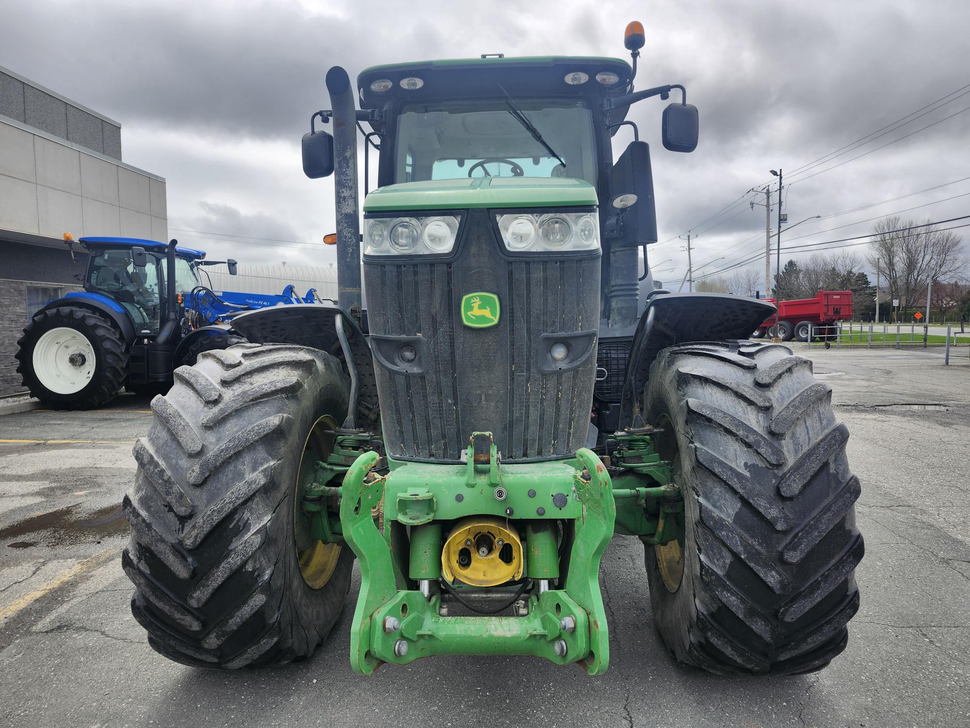 Vue de face d'un tracteur John Deere vert garé sur le trottoir, avec ses gros pneus robustes et son extérieur sale sous un ciel nuageux. Un autre tracteur bleu, un camion rouge et des bâtiments apparaissent en arrière-plan.
