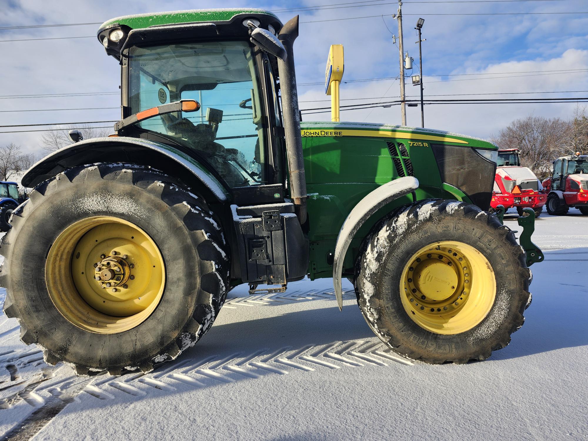 Un tracteur vert avec de gros pneus est garé sur une surface enneigée avec des traces de pneus visibles. Le ciel est partiellement nuageux et des lignes électriques sont visibles en arrière-plan.