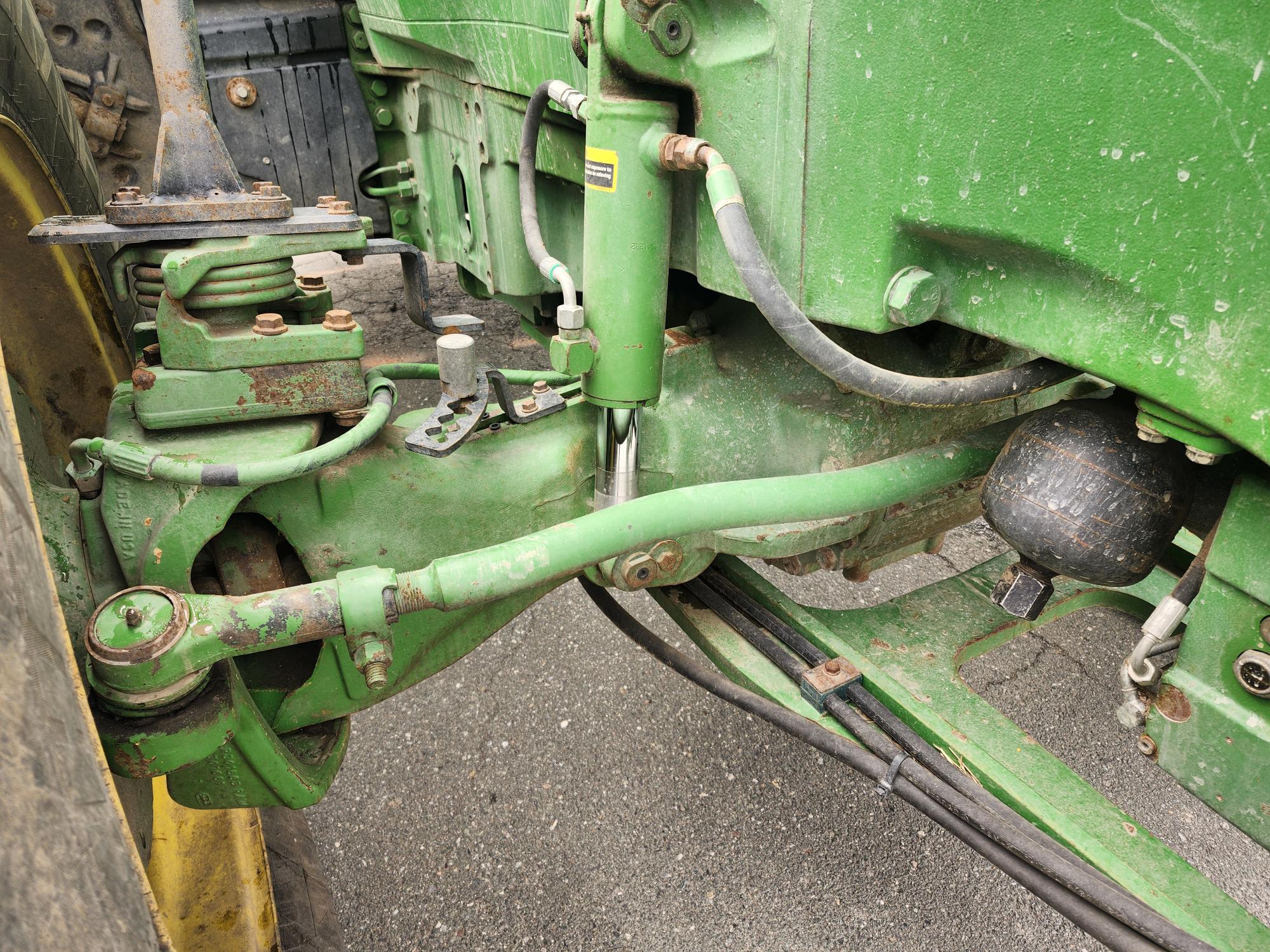 Gros plan de l'essieu avant et du système de suspension d'un tracteur vert, montrant des conduites hydrauliques, des tiges métalliques, des joints et un peu de rouille, avec une partie d'un gros pneu visible sur la gauche.