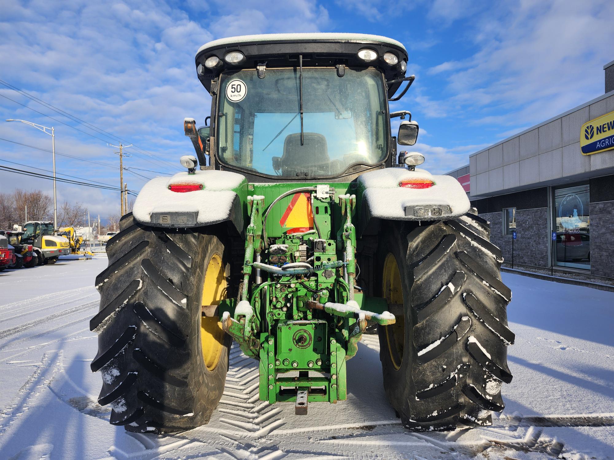 Un tracteur vert recouvert de plaques de neige est stationné sur une surface enneigée. Le tracteur est équipé de gros pneus noirs et d'un mécanisme d'attache arrière. À l'arrière-plan, on voit un bâtiment et un ciel bleu clair.