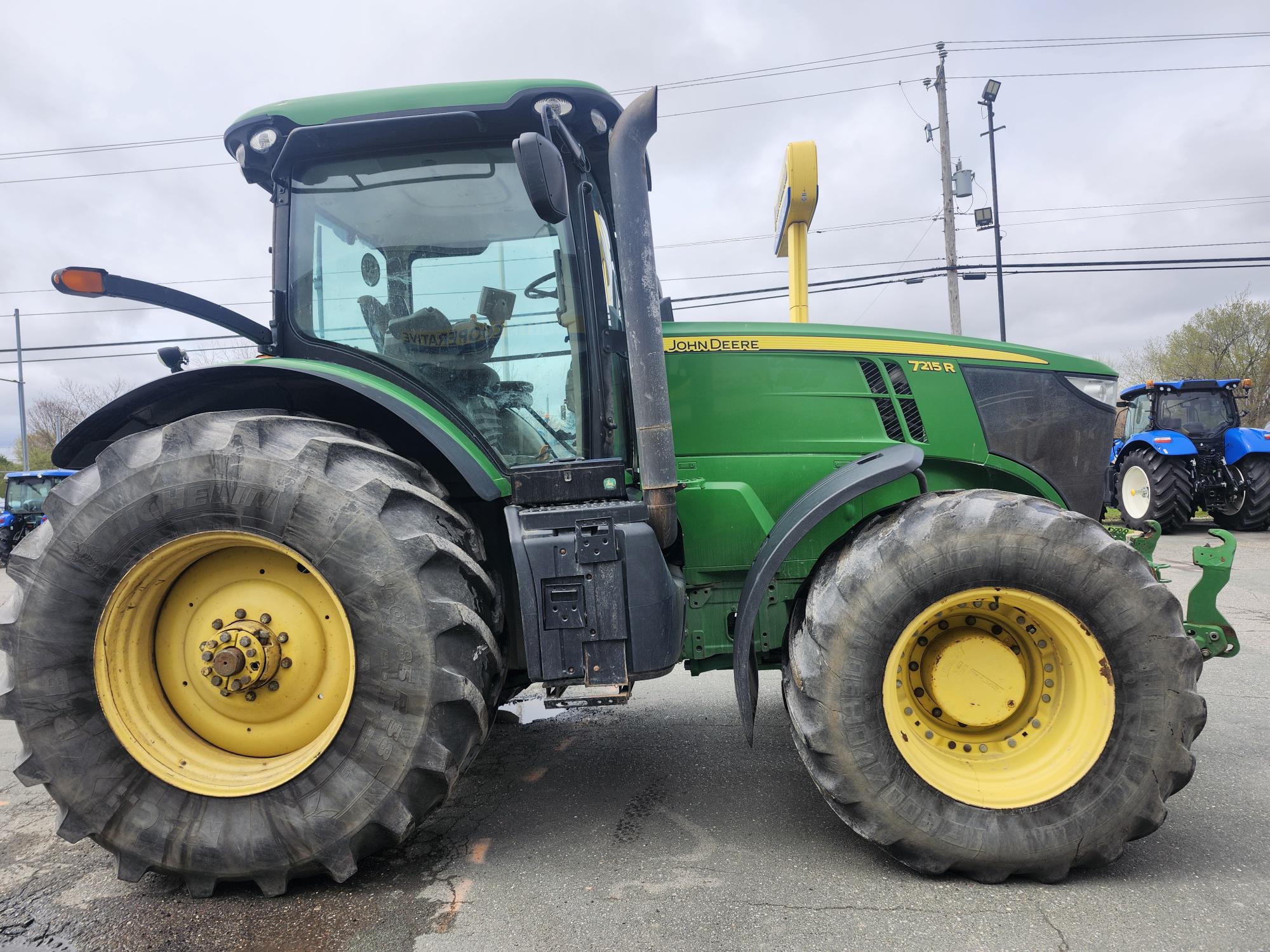 Un tracteur John Deere 7215R vert avec de grandes roues jaunes est garé sur le trottoir. Des lignes électriques et un tracteur bleu sont visibles en arrière-plan sous un ciel nuageux.