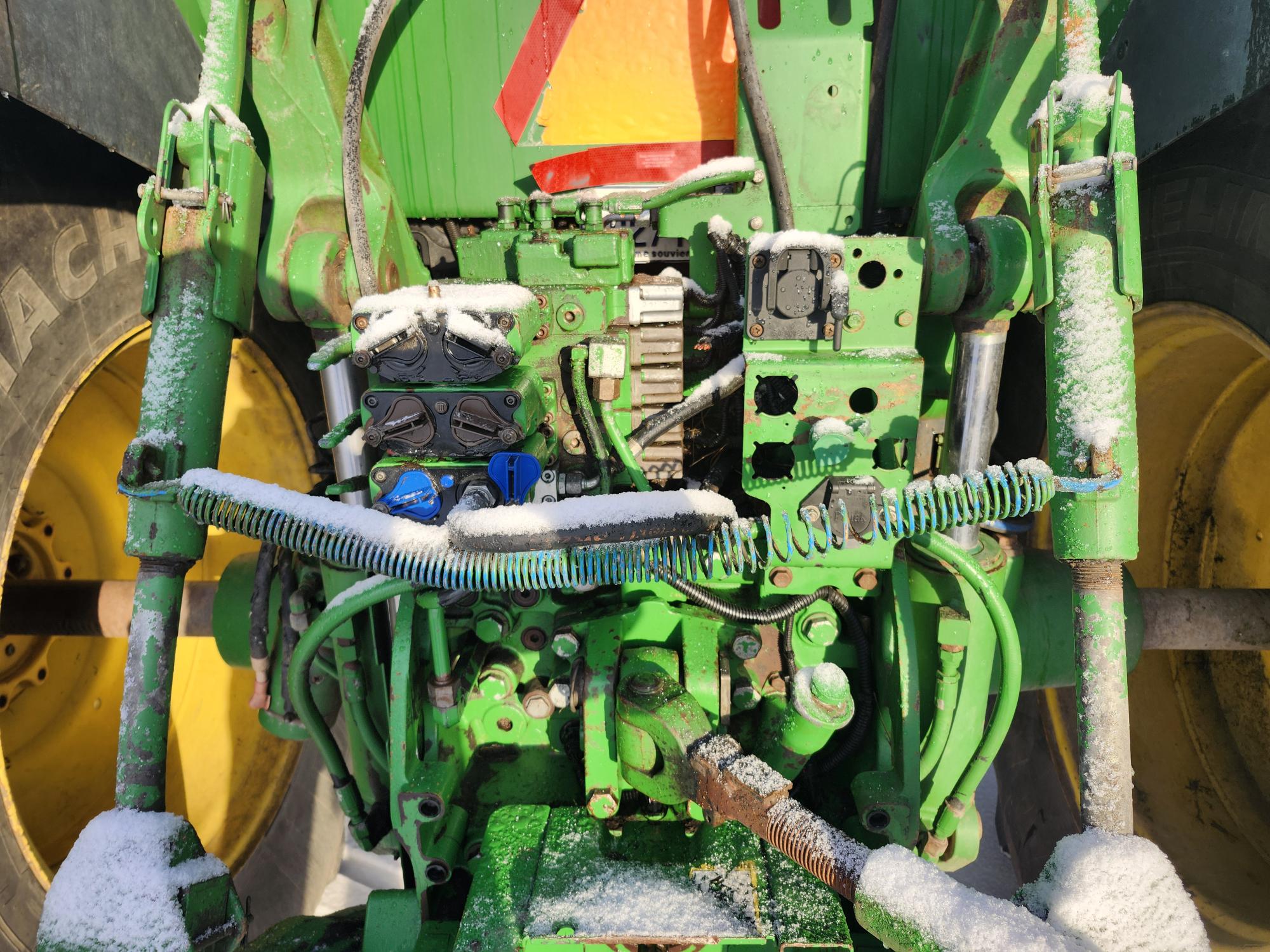 Gros plan sur les connexions mécaniques arrière d'un tracteur vert, saupoudré de neige. La machine est dotée de divers câbles et composants avec un gros pneu visible de chaque côté. Un triangle réfléchissant rouge est partiellement visible au-dessus.