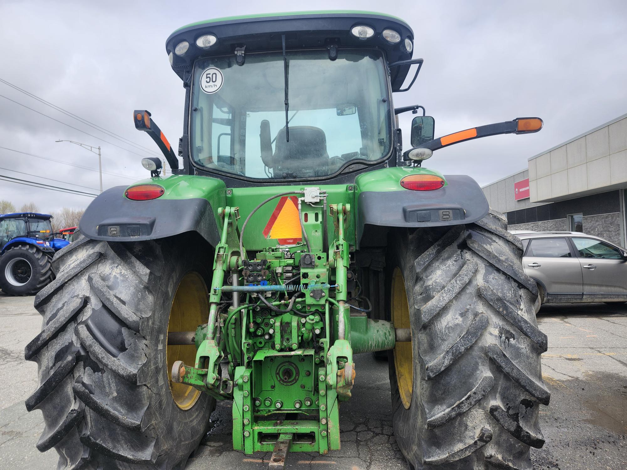 Un tracteur vert vu de l'arrière, montrant de gros pneus arrière, un panneau triangulaire indiquant un véhicule lent et des composants mécaniques ; garé à l'extérieur près de voitures et d'un autre tracteur par temps nuageux.