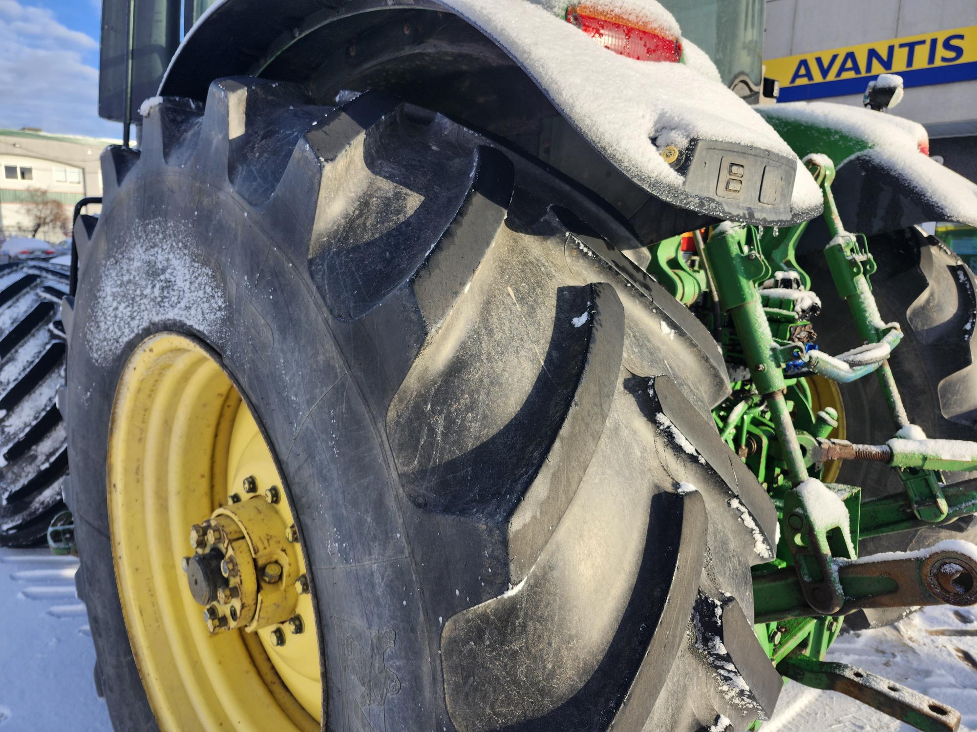 Gros plan d'un pneu de tracteur recouvert de neige, avec une partie du tracteur et de sa machinerie verte visibles. L'arrière-plan montre un bâtiment sur lequel est inscrit le mot 