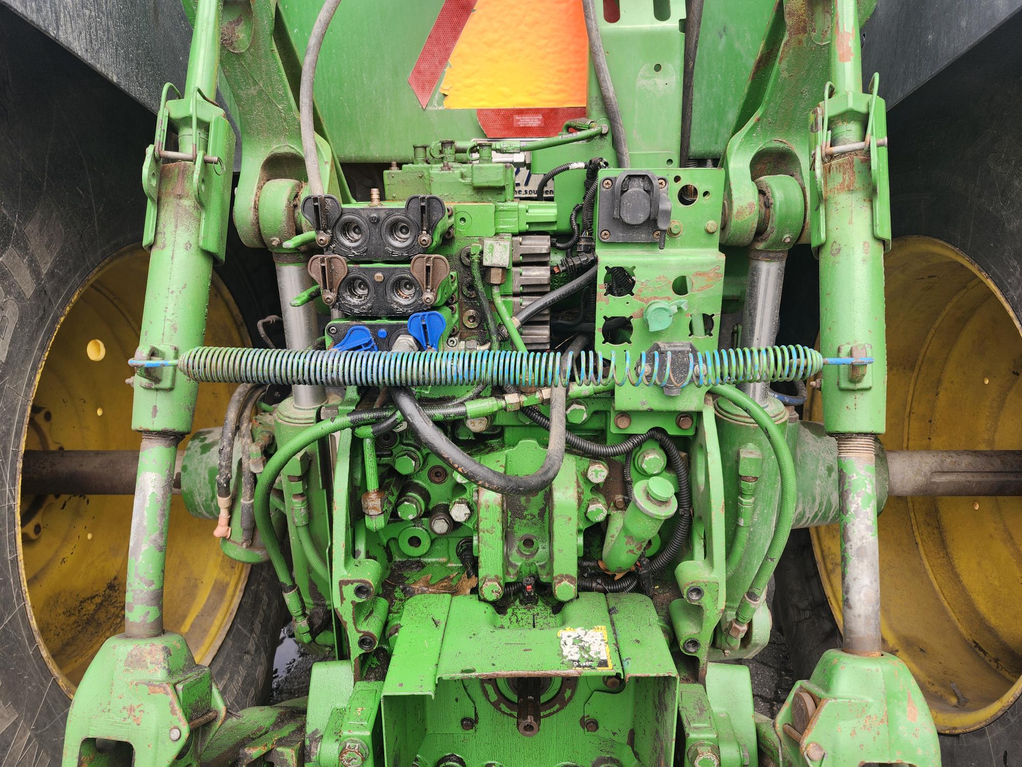 Gros plan du système hydraulique arrière et de l'attelage d'un tracteur vert, montrant divers tuyaux, leviers et composants métalliques, avec de grandes roues jaunes visibles des deux côtés.