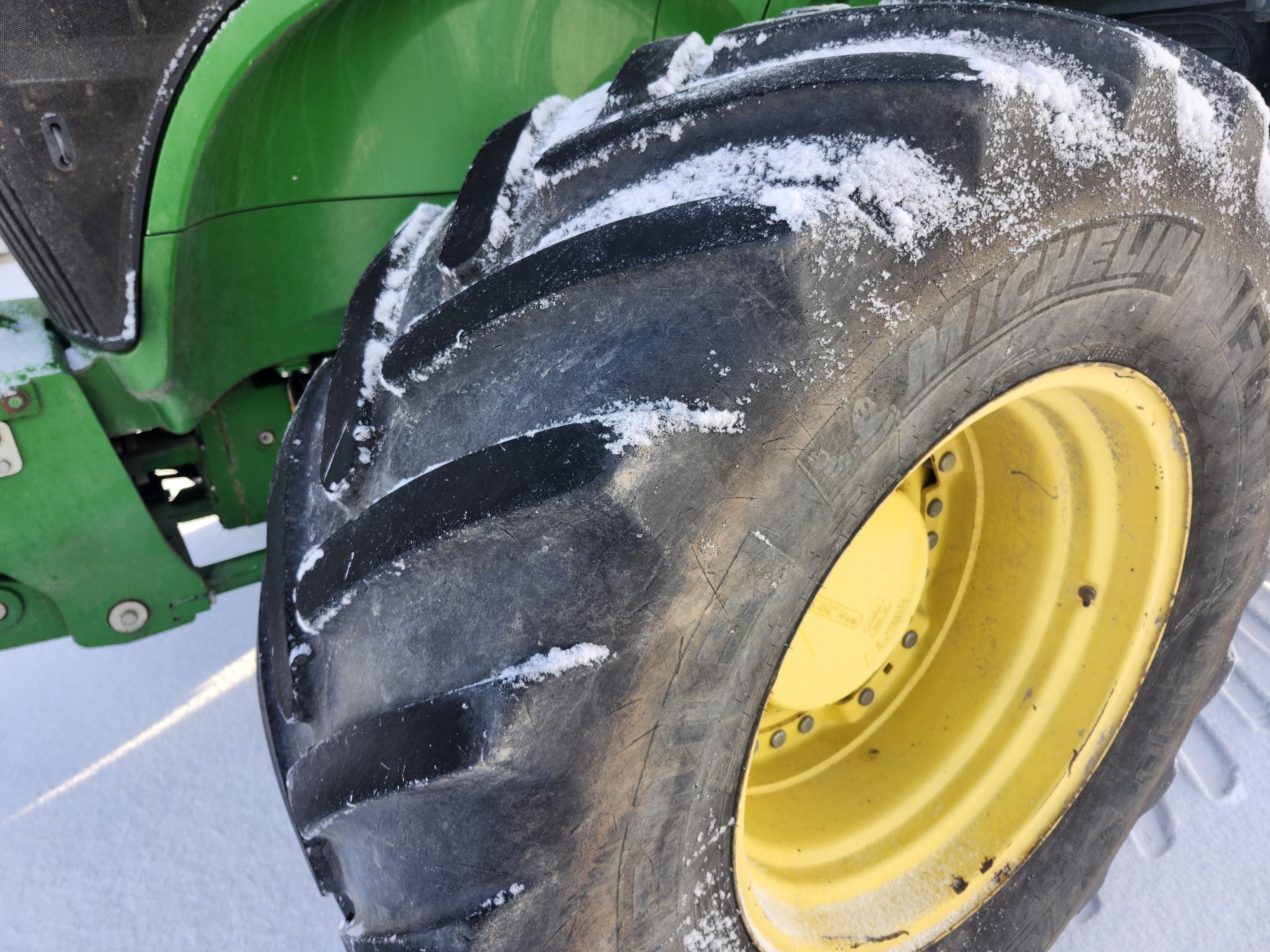 Gros plan d'un pneu de tracteur avec de la neige visible dans les bandes de roulement. Le pneu est recouvert de plaques de neige et la carrosserie verte du tracteur est partiellement visible à l'arrière-plan. Le sol est recouvert de neige.