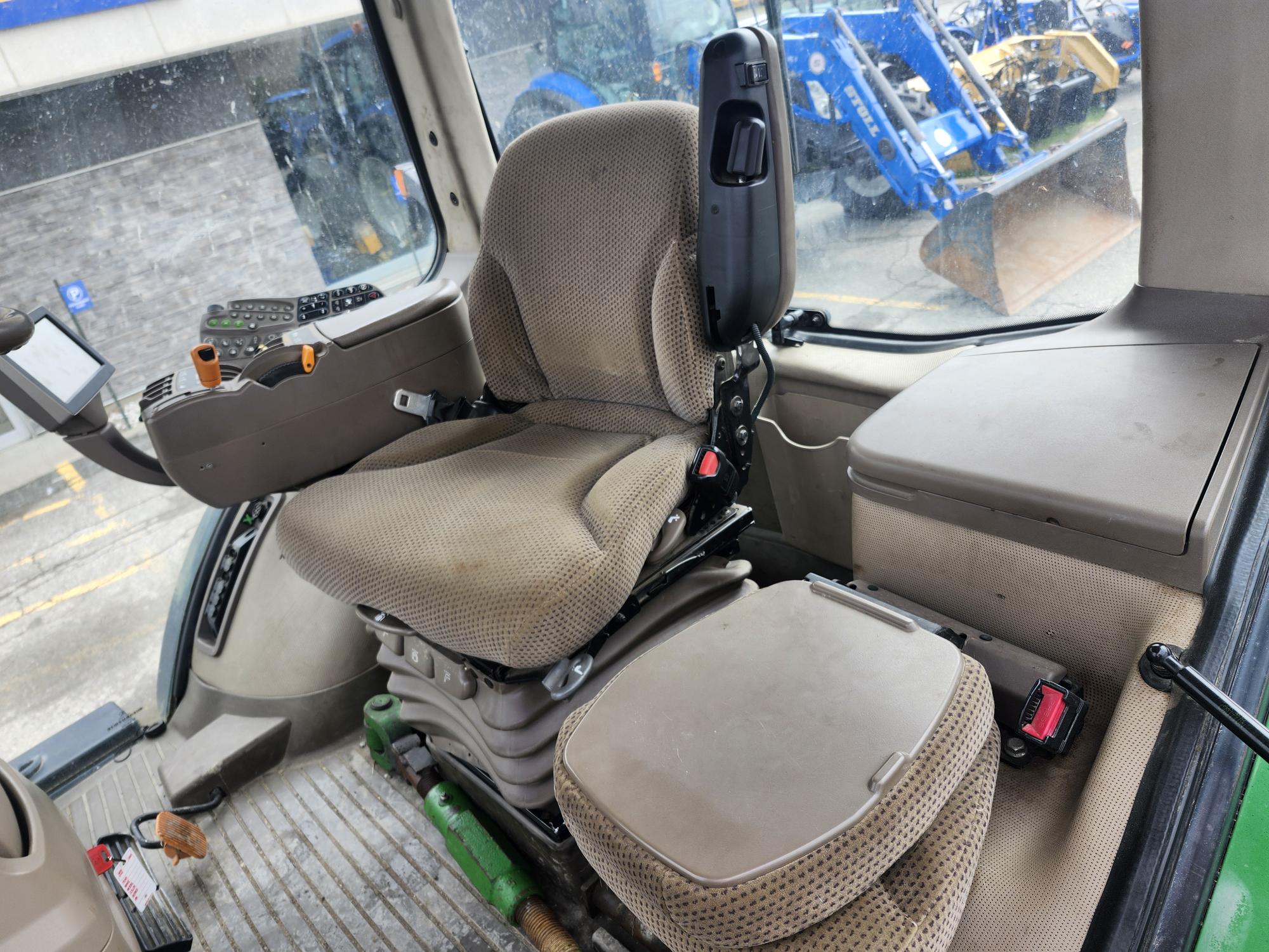L'intérieur d'une cabine de tracteur comprenant un siège conducteur marron rembourré avec accoudoirs, des panneaux de commande à gauche et un siège d'appoint plus petit à côté ; à l'extérieur, des tracteurs bleus sont visibles à travers les fenêtres.