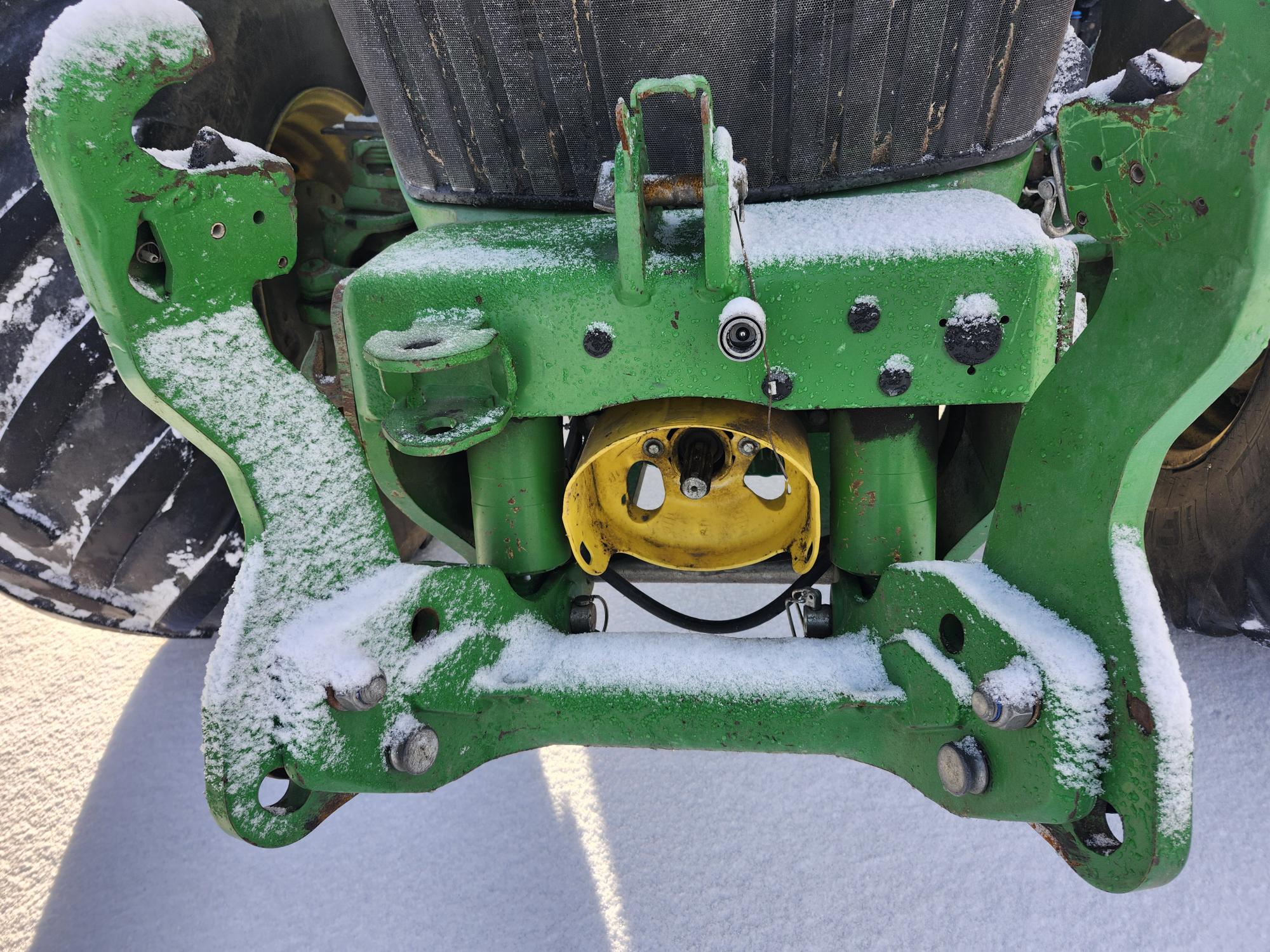 Gros plan de la partie avant d'un tracteur vert, recouverte d'une fine couche de neige. Le point d'attache central jaune du tracteur et les gros pneus noirs sont visibles. La surface métallique présente des signes d'usure et de vieillissement.