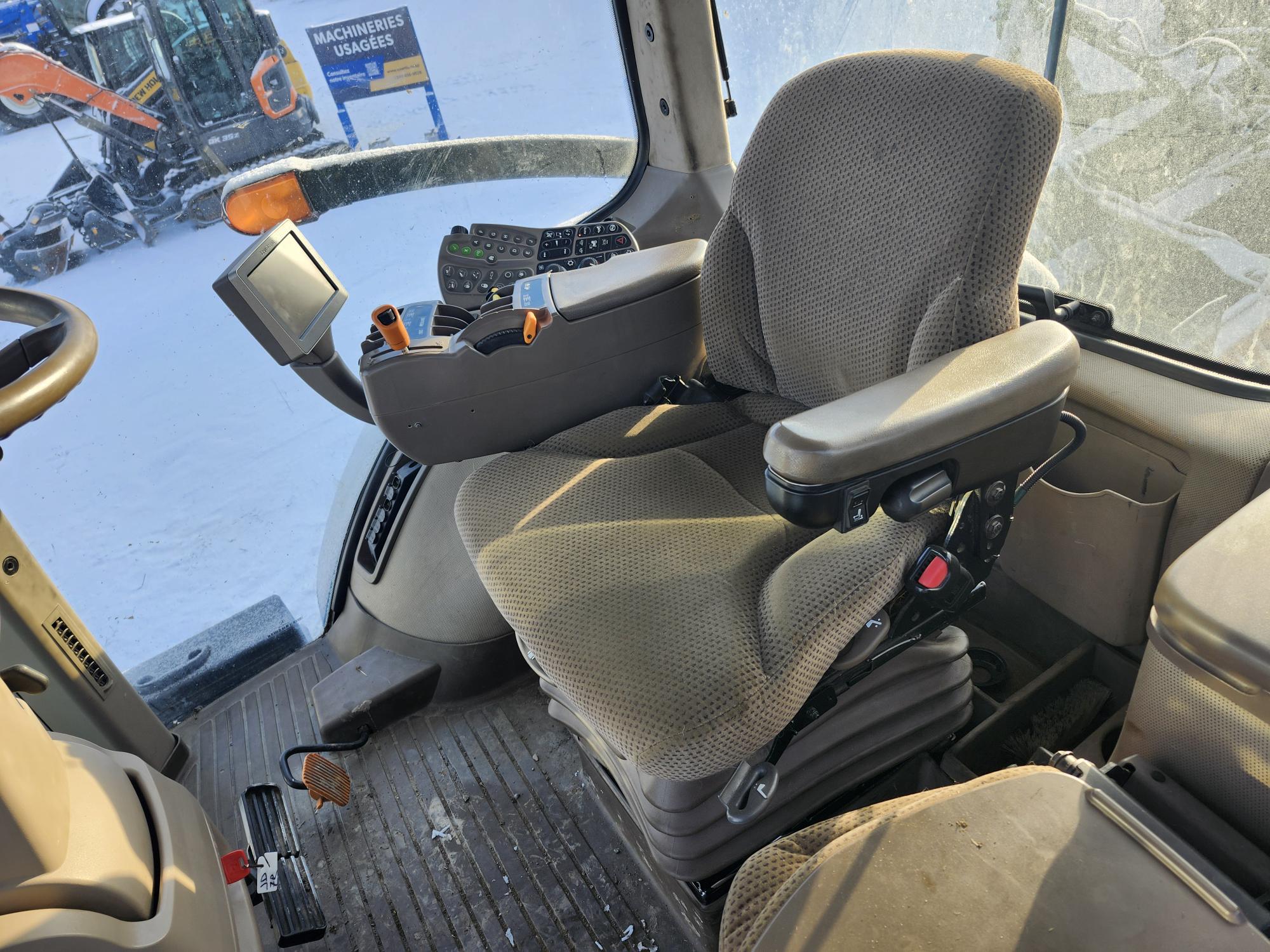 Intérieur d'une cabine de véhicule avec siège conducteur, panneau de commande et volant recouverts de tissu. La lumière du soleil traverse les fenêtres, éclairant l'équipement. De la neige et des engins de chantier sont visibles à l'extérieur.