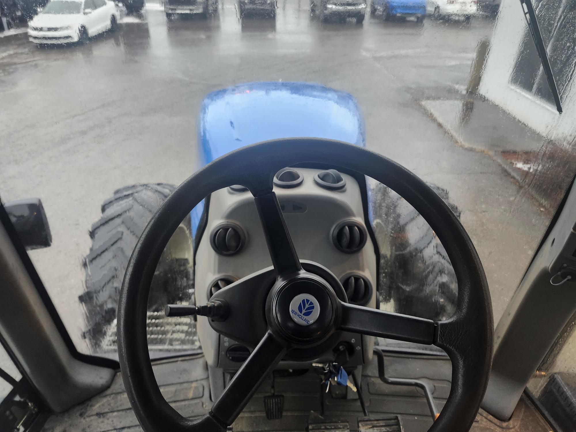 Vue de l'intérieur de la cabine d'un tracteur, montrant le volant et les commandes du tableau de bord. Le pare-brise avant est légèrement flou à cause de la pluie. À l'extérieur, plusieurs véhicules sont stationnés sur une surface mouillée.