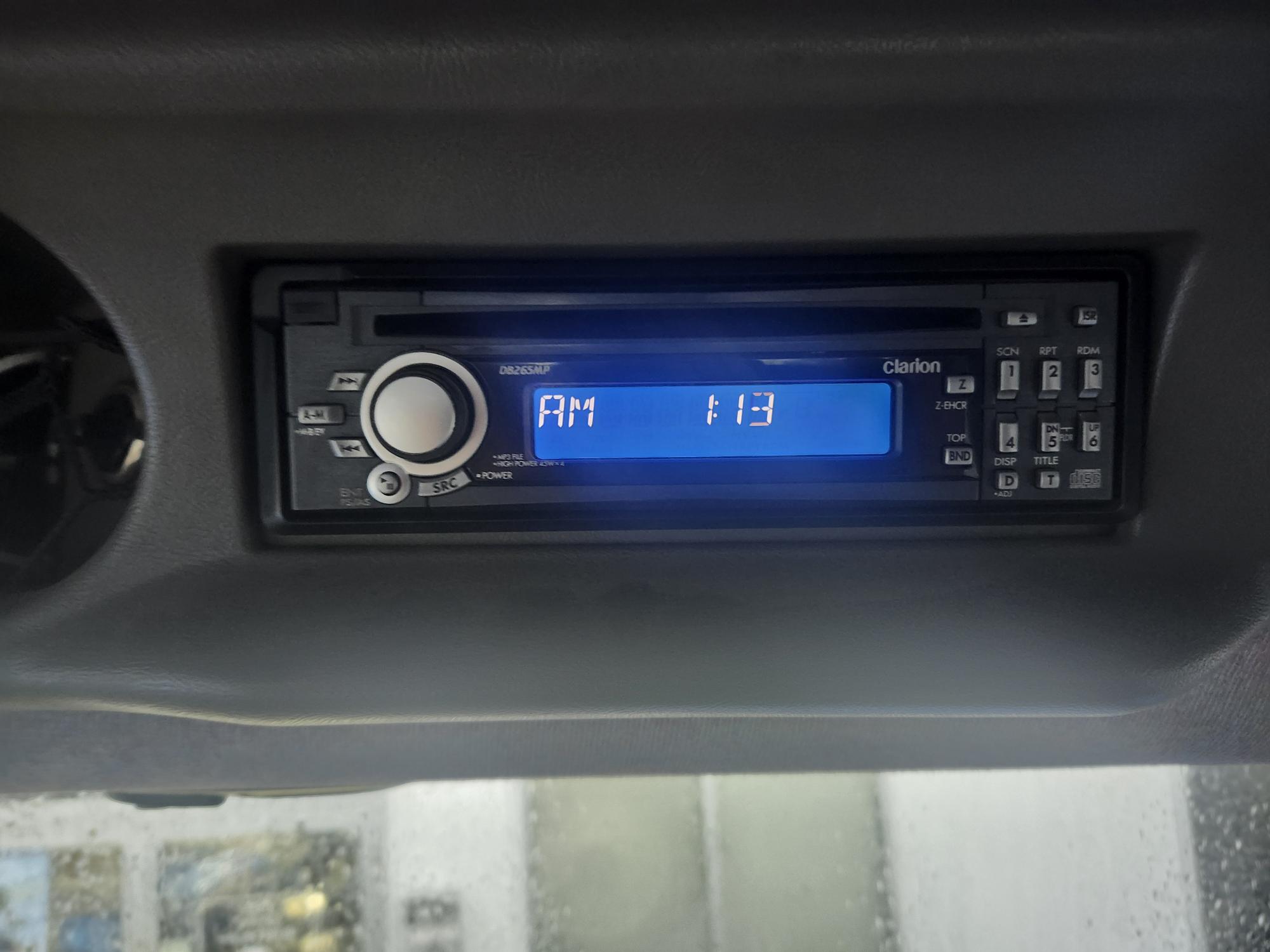 Autoradio avec écran numérique rétroéclairé bleu indiquant « AM 1:13 ». L'appareil est doté de plusieurs boutons et d'un bouton rotatif à gauche. Il est installé dans le tableau de bord d'un véhicule.