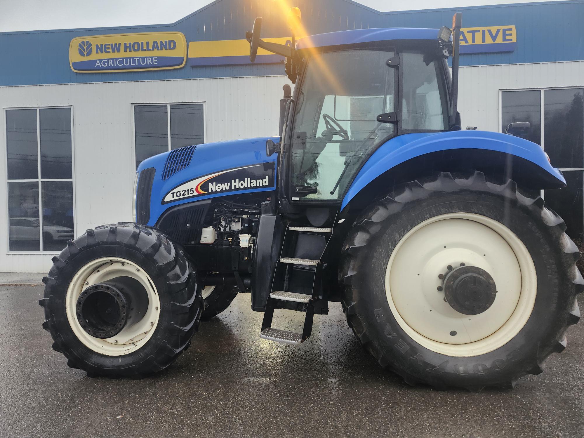 Tracteur New Holland TG215 bleu stationné sur une chaussée mouillée devant un bâtiment portant une pancarte New Holland Agriculture. Le tracteur est grand, avec de gros pneus, et est vu de côté.