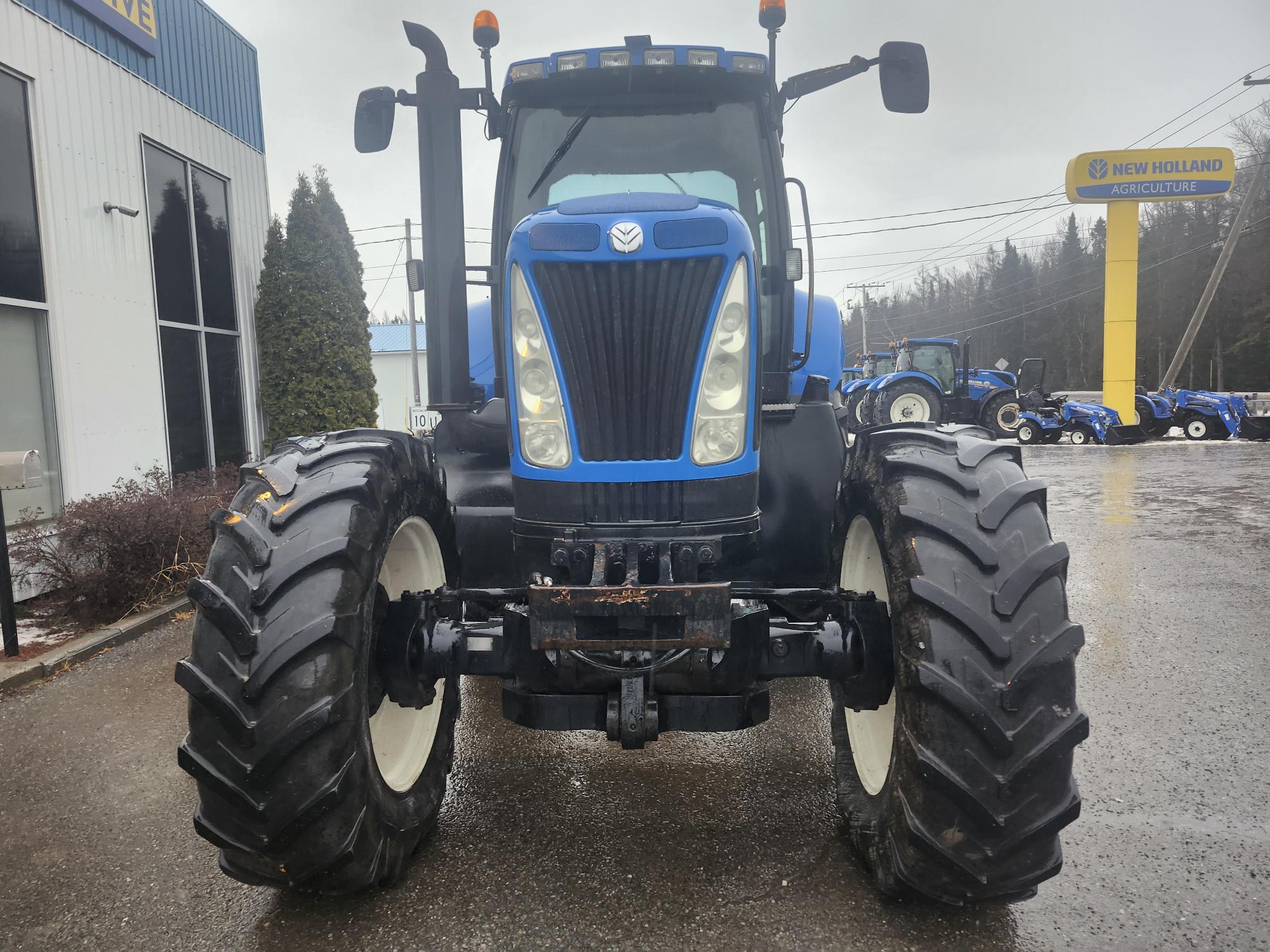 Un tracteur New Holland bleu est garé sur une surface mouillée à l'extérieur d'un concessionnaire, avec d'autres tracteurs en arrière-plan et un panneau jaune New Holland Agriculture visible à droite. Le ciel est couvert, ce qui suggère un temps pluvieux.