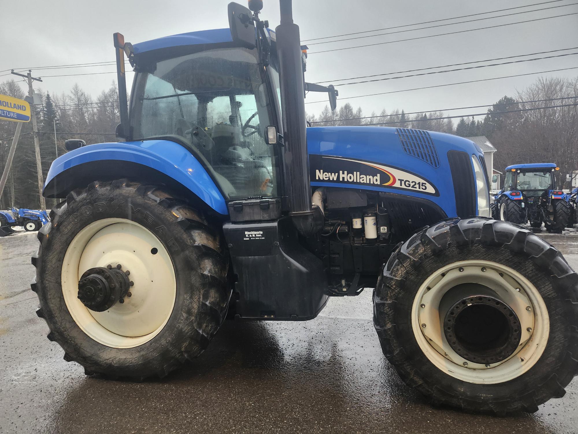 Un tracteur New Holland TG215 bleu est garé sur une surface mouillée par temps nuageux. Le panneau de New Holland est visible en arrière-plan, ainsi qu'un autre tracteur et quelques arbres.