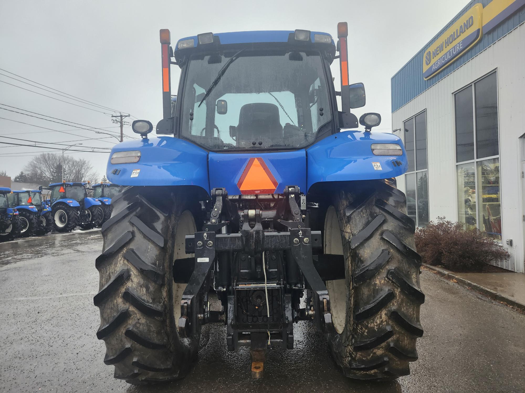 Un tracteur bleu vu de l'arrière, stationné sur une chaussée mouillée. Il est équipé de gros pneus et d'un panneau réfléchissant triangulaire. Plusieurs autres tracteurs et un bâtiment portant l'inscription « New Holland » sont visibles en arrière-plan.