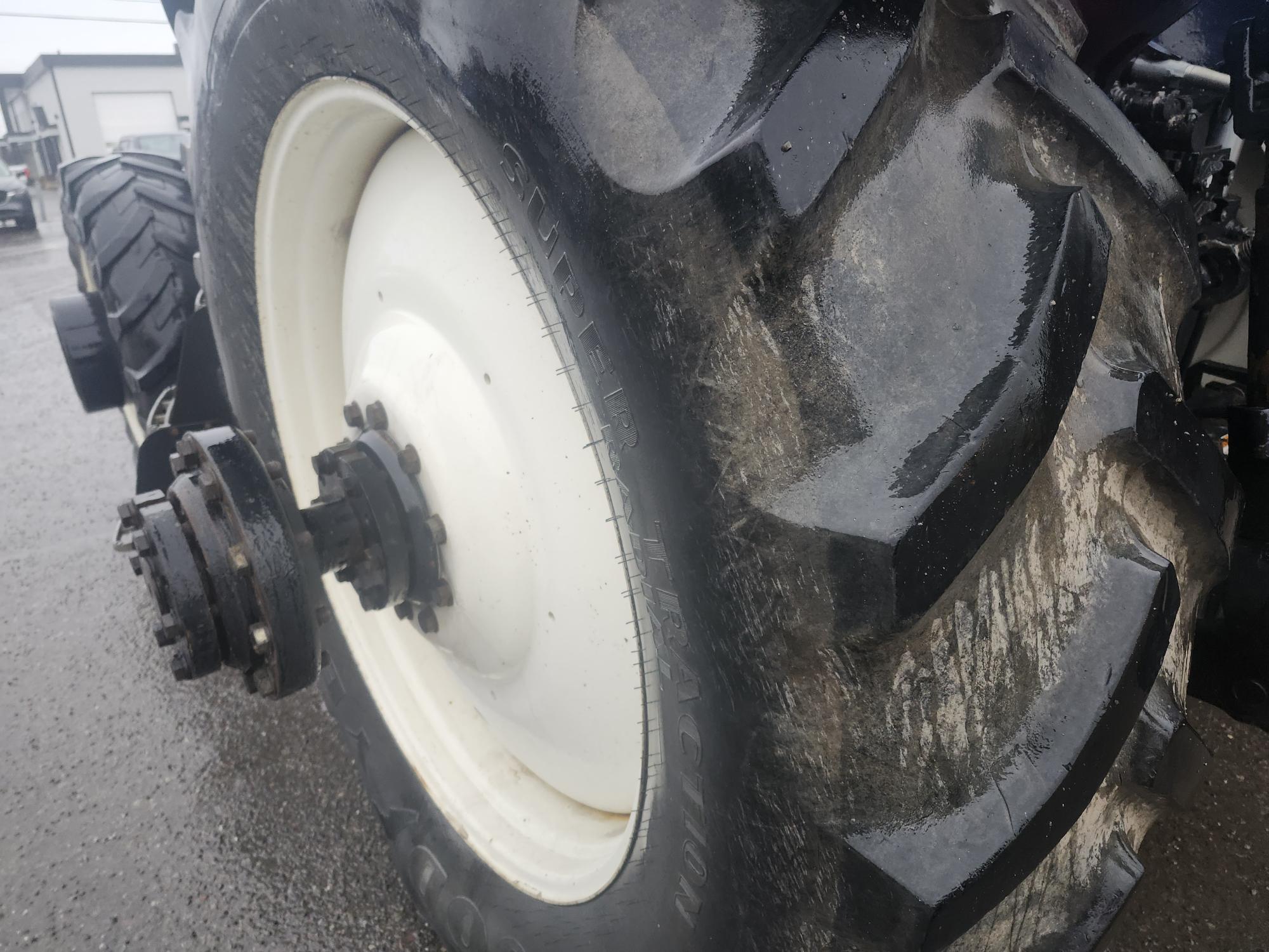 Gros plan d'un pneu de tracteur boueux sur une surface pavée et mouillée. Les profondes rainures du pneu sont visibles et il y a un autre pneu en arrière-plan. Le décor semble couvert et industriel.