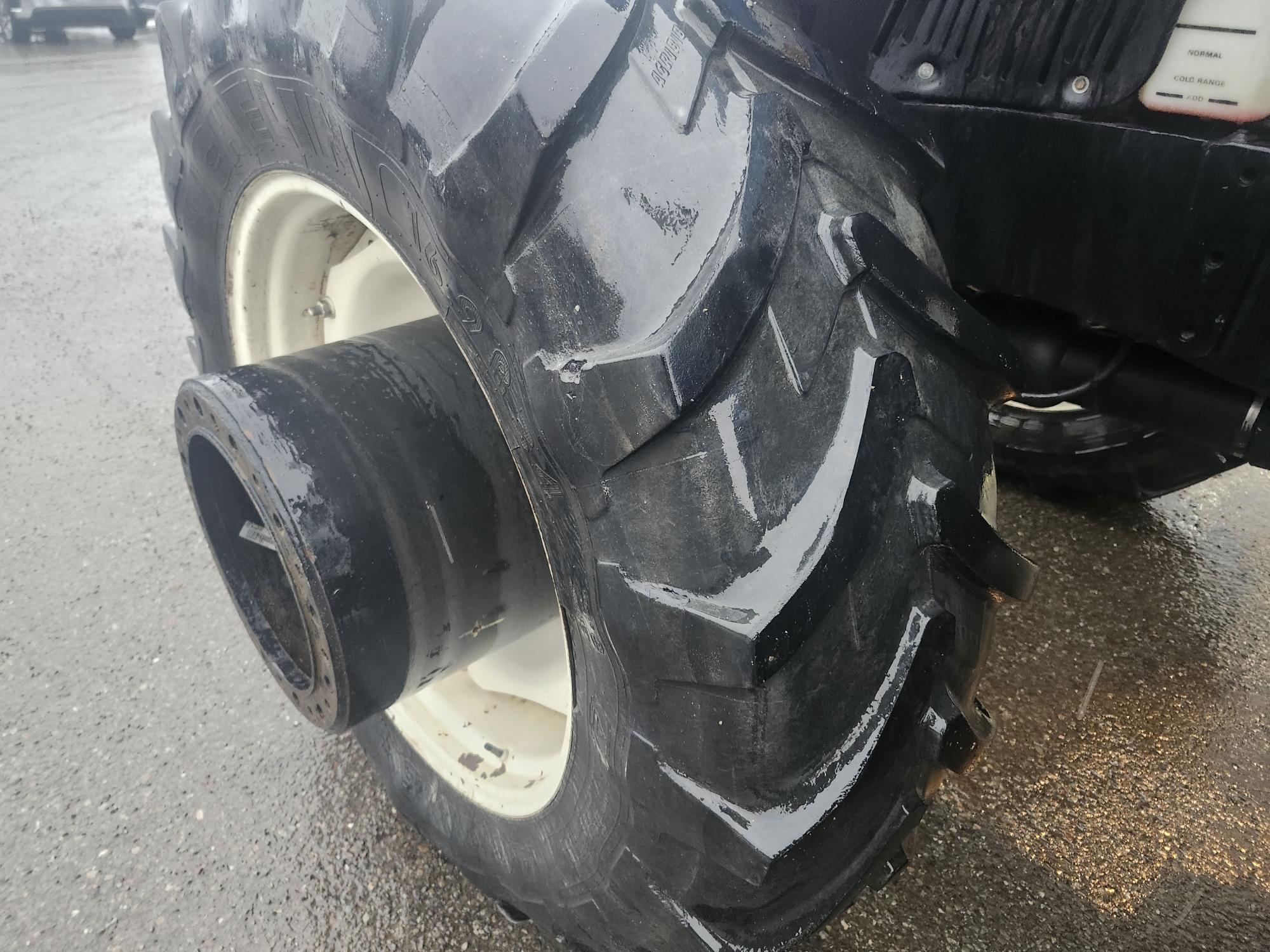 Gros plan d'un gros pneu de tracteur robuste sur une surface pavée et mouillée. La bande de roulement du pneu est profonde et conçue pour une utilisation intensive, ce qui suggère qu'il fait partie d'une machine agricole ou de construction.