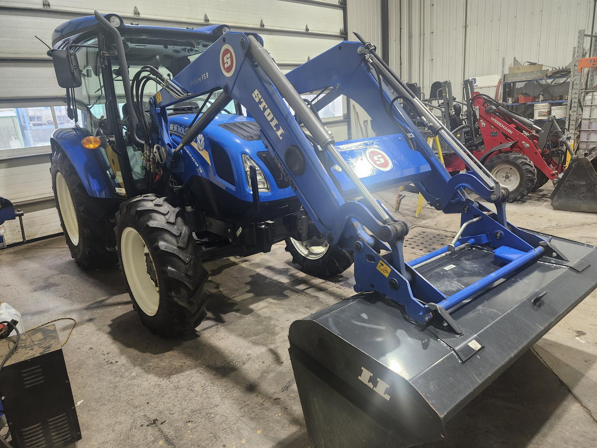 Un tracteur bleu équipé d'un chargeur frontal est stationné dans un garage. Le chargeur est relevé et divers outils et équipements sont visibles en arrière-plan.