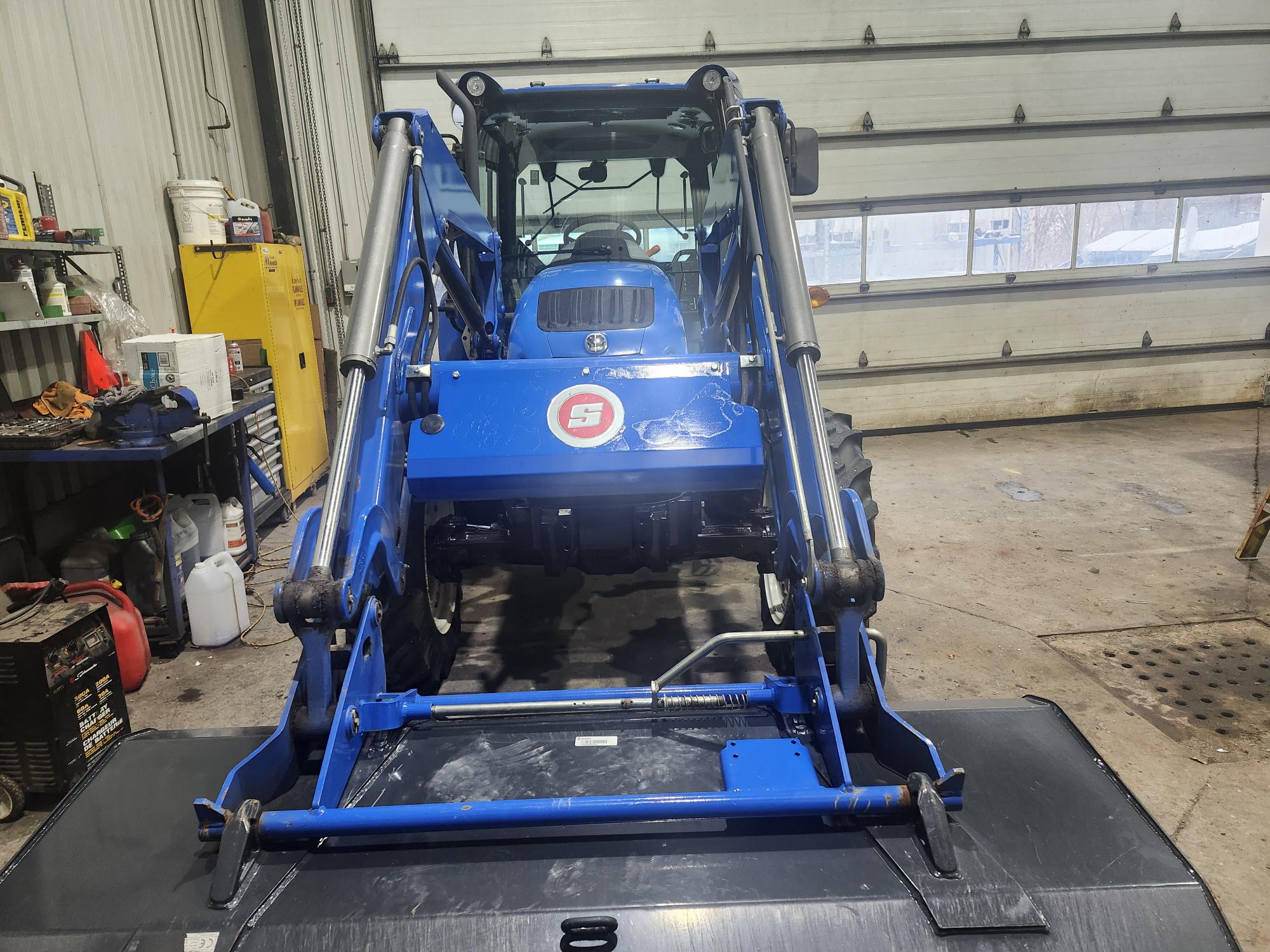 Un tracteur bleu équipé d'un chargeur frontal est garé dans un garage. Le garage est doté de grandes portes, d'étagères avec divers articles et d'équipements disposés tout autour. La scène suggère une zone d'entretien ou de stockage pour le tracteur.