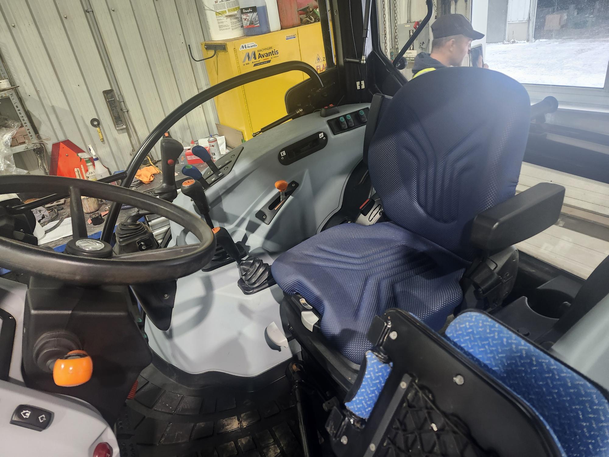 L'image montre l'intérieur d'une cabine de tracteur avec un siège conducteur, un volant et des leviers de commande. La cabine est moderne, avec une sellerie bleue et diverses commandes sur le tableau de bord. Une personne est visible à l'arrière-plan à l'extérieur de la cabine.