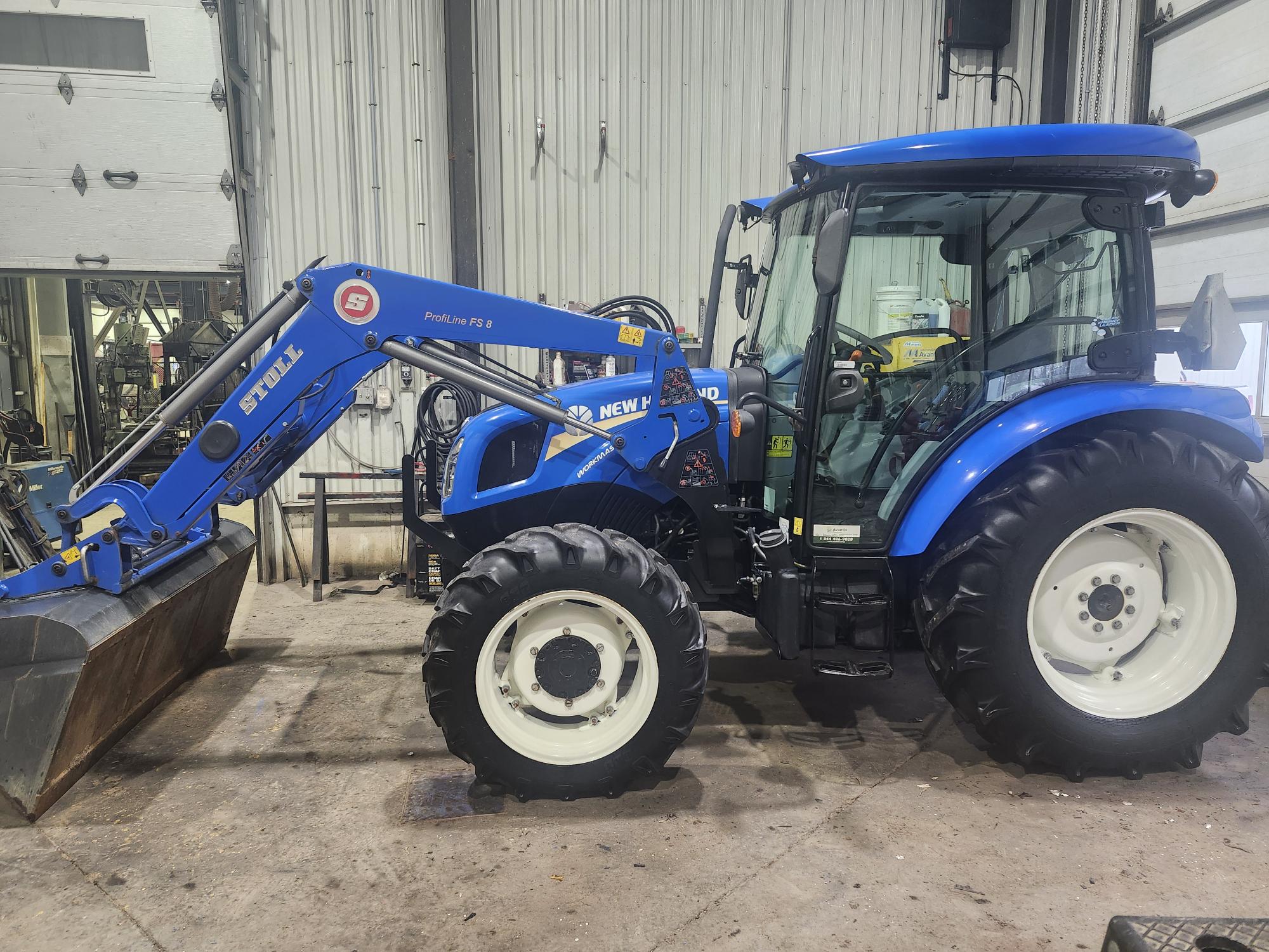 Un tracteur New Holland bleu équipé d'un chargeur frontal est garé à l'intérieur d'un garage. La zone est bien éclairée avec des murs blancs et divers outils visibles en arrière-plan.