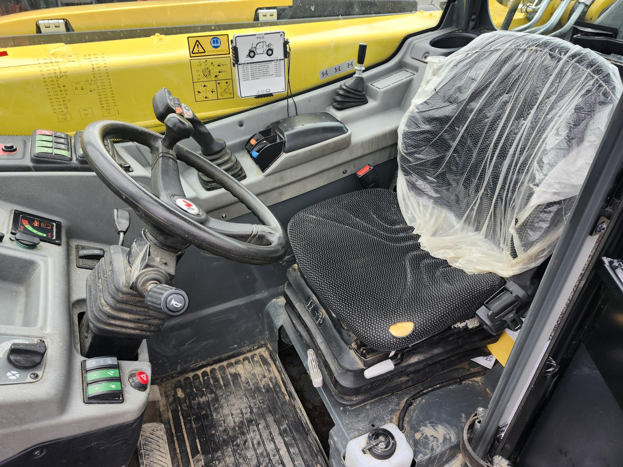 La cabine intérieure d'un véhicule de construction, montrant un volant, divers leviers et boutons de commande, un siège opérateur noir recouvert de plastique et des étiquettes d'instructions de sécurité jaunes sur le tableau de bord.