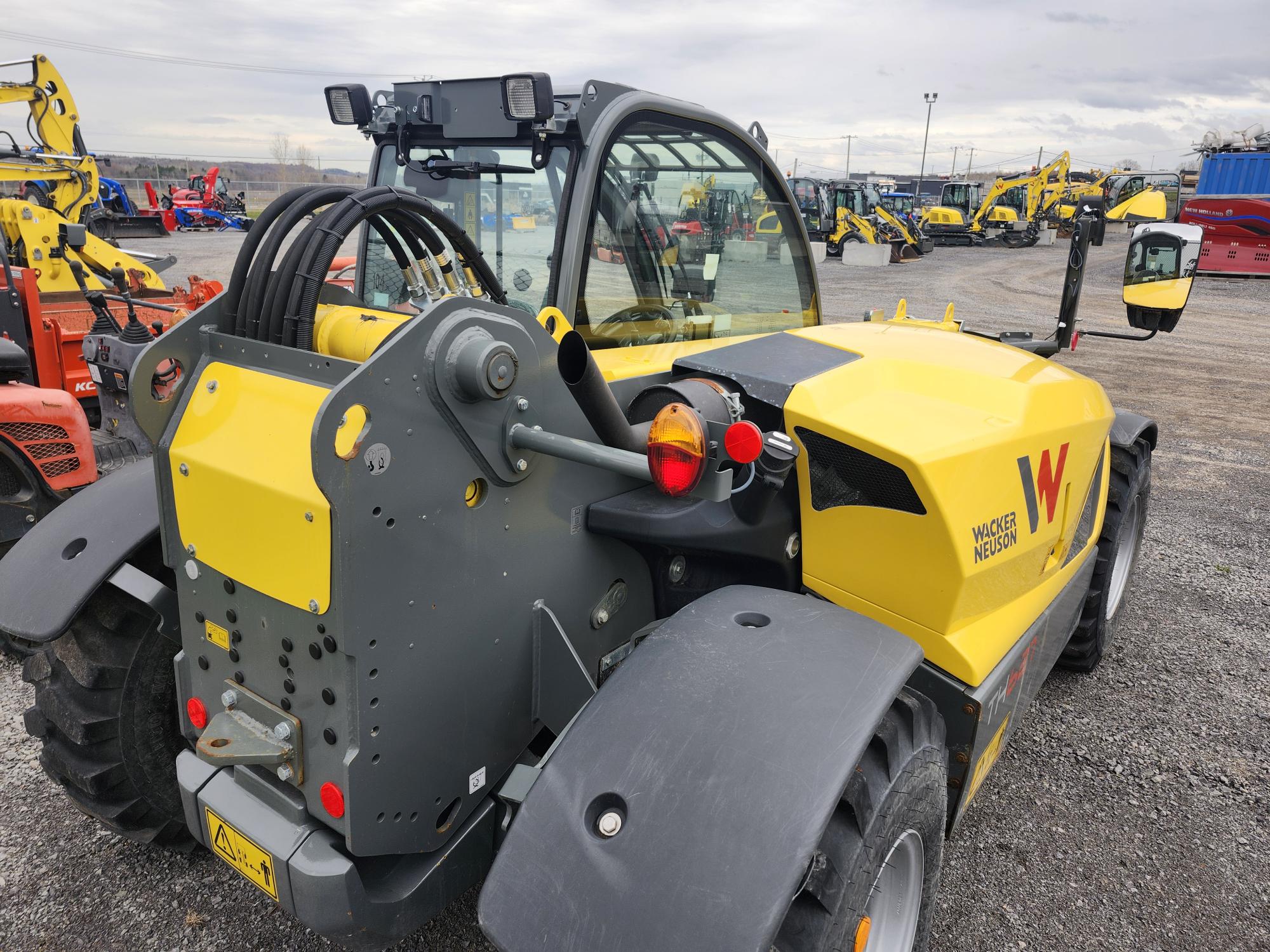 Un chariot télescopique Wacker Neuson jaune est garé sur un terrain en gravier, parmi divers véhicules et engins de chantier. La photo montre une vue arrière et latérale, mettant en valeur la cabine, les pneus et les composants hydrauliques de l'engin.