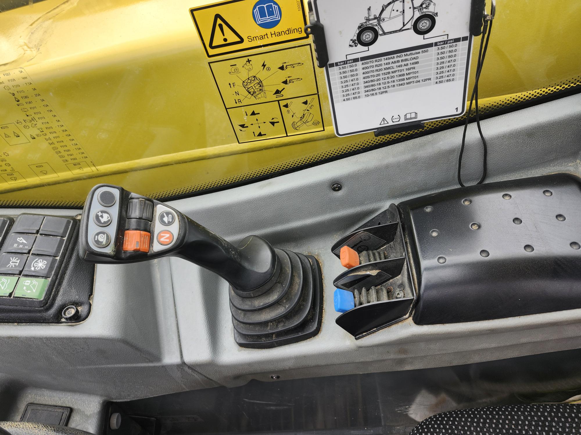 Gros plan d'un panneau de commande à l'intérieur d'une cabine de véhicule ou de machine, montrant un joystick avec des boutons, des leviers avec des pointes bleues et oranges, des autocollants d'avertissement et un schéma pour les instructions d'utilisation.
