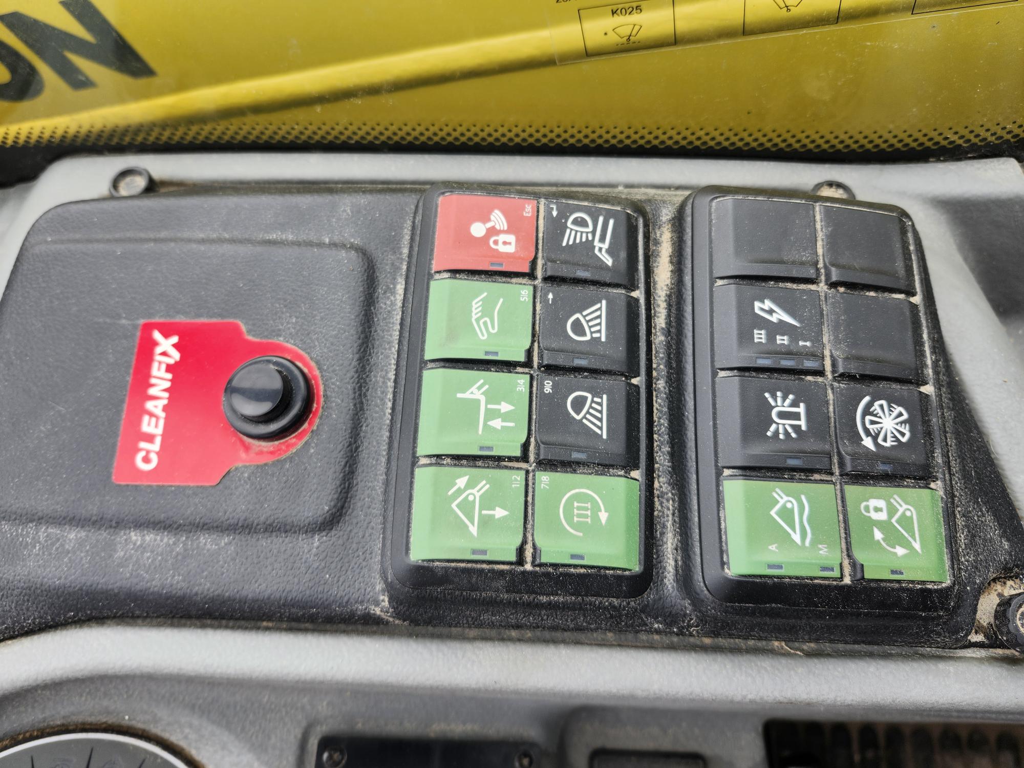 Gros plan sur le panneau de commande d'un véhicule avec divers boutons, dont des boutons verts et noirs pour différentes fonctions, et un bouton rouge portant l'inscription « CLEANFIX ». Une étiquette d'avertissement jaune est partiellement visible au-dessus.