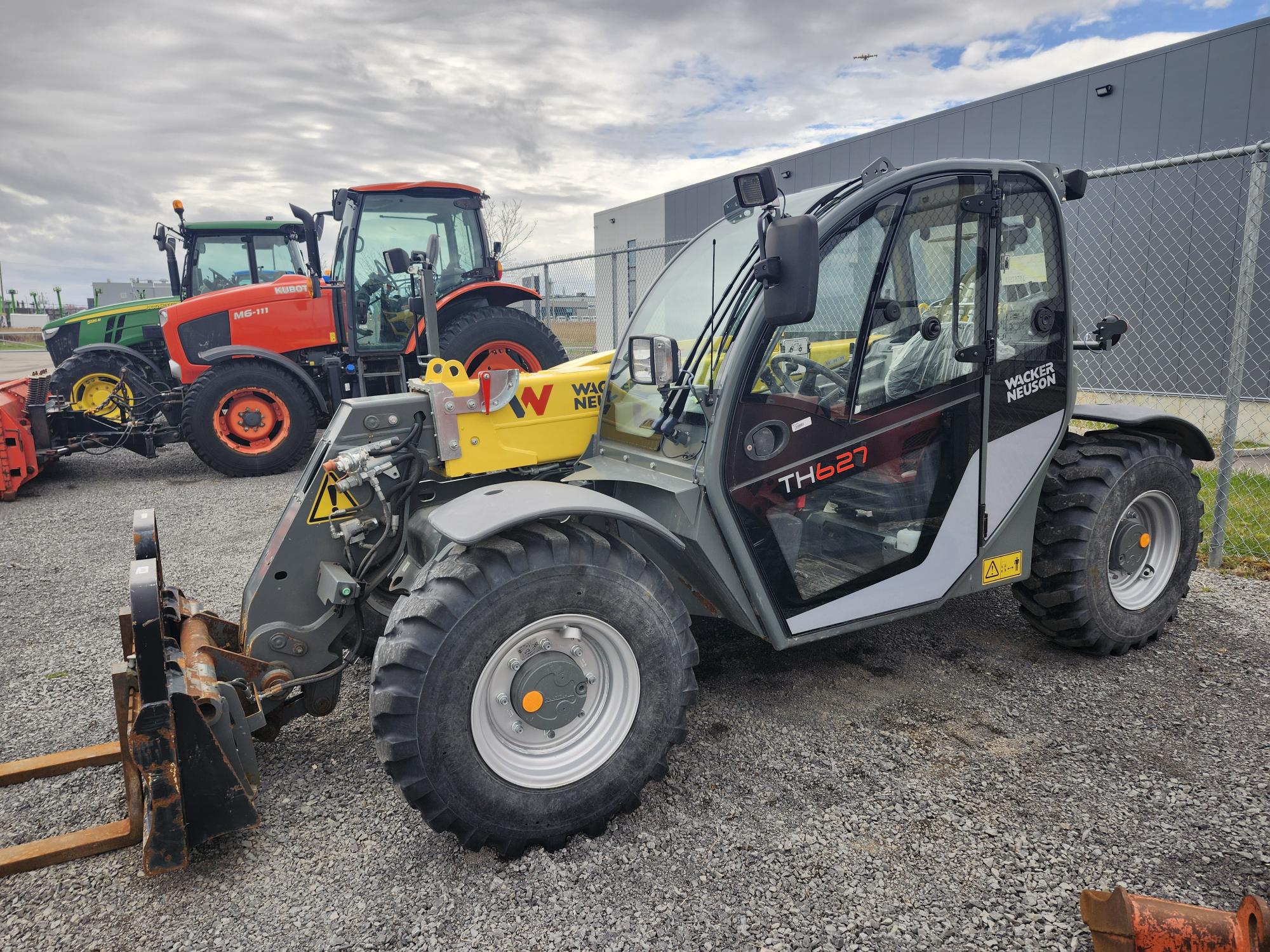Un chariot télescopique Wacker Neuson TH627 est garé sur du gravier près d'une clôture grillagée, avec un tracteur orange et un bâtiment gris visibles en arrière-plan sous un ciel nuageux.