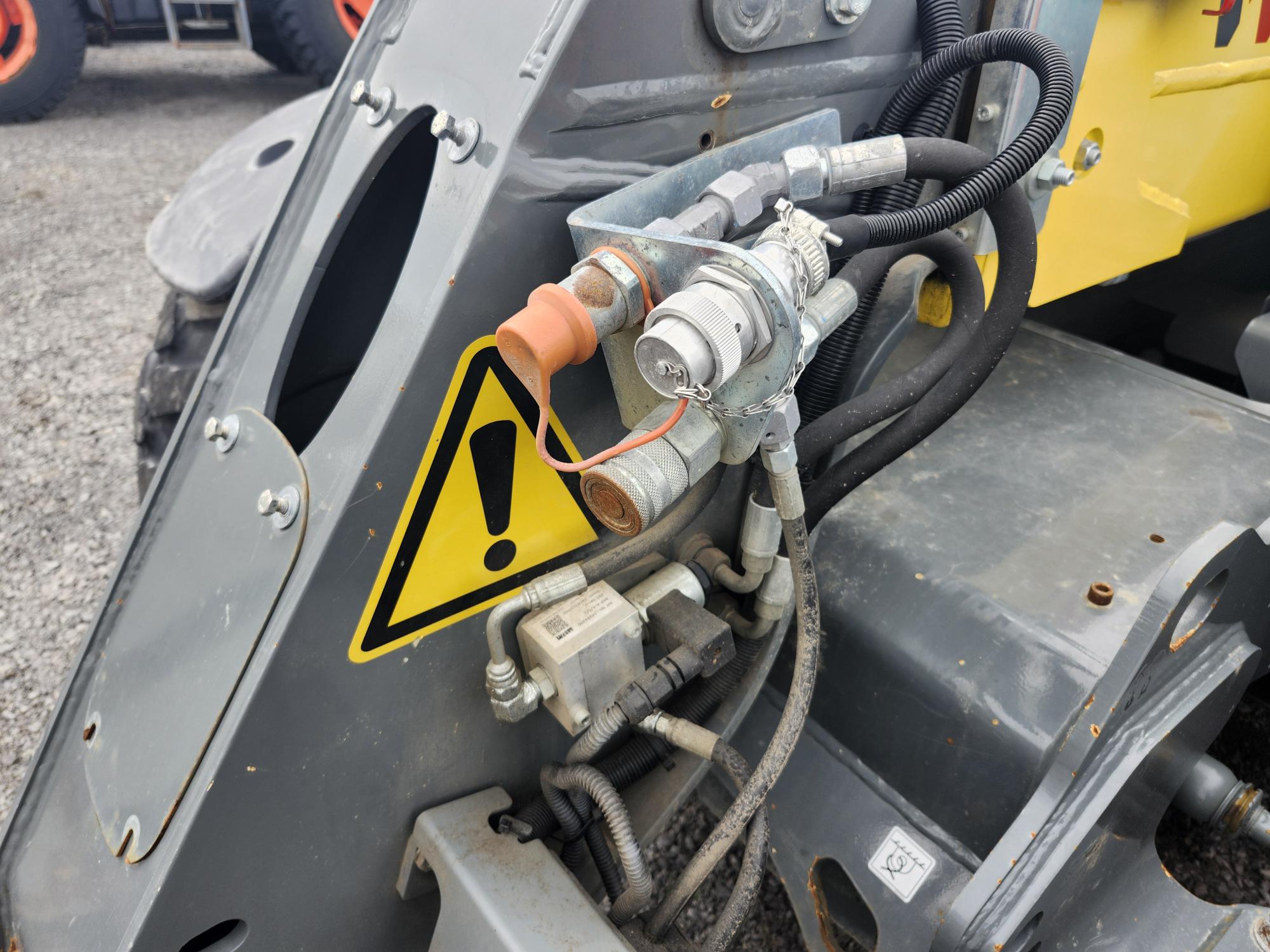 Gros plan sur une machine industrielle montrant des flexibles hydrauliques, des raccords métalliques et des câbles à côté d'un panneau d'avertissement jaune avec un point d'exclamation noir sur une surface métallique grise. Un sol en gravier est visible en arrière-plan.