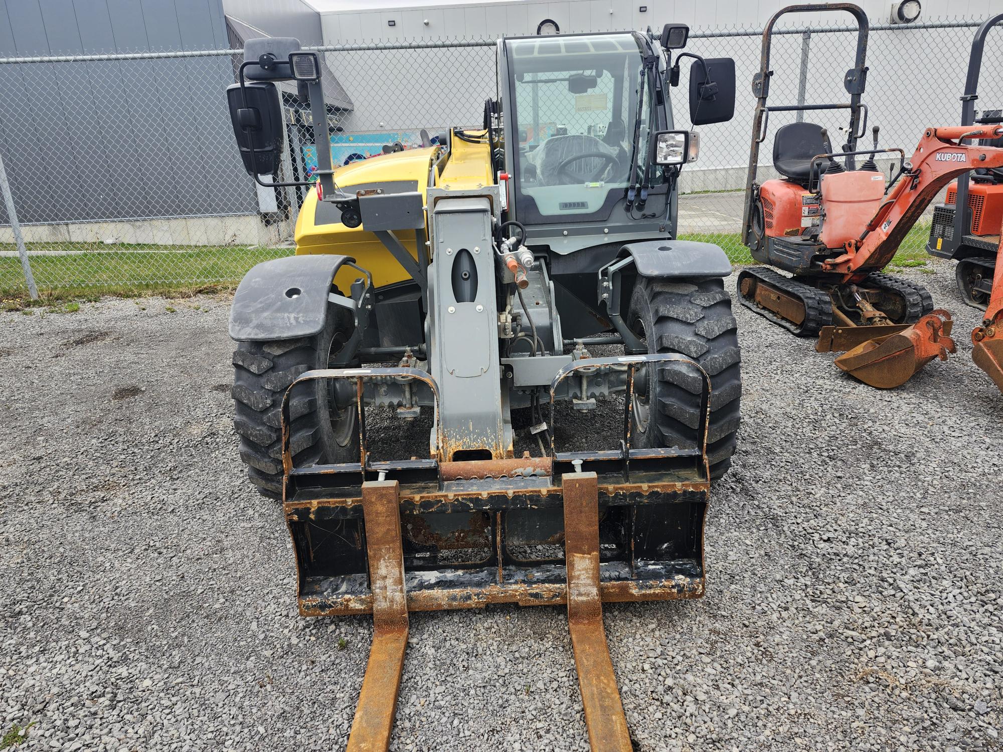 Un chariot télescopique jaune et gris à gros pneus est garé sur du gravier, face à la caméra. Son attache-fourche rouillée est bien visible au premier plan. Deux autres engins de chantier orange sont visibles à droite.
