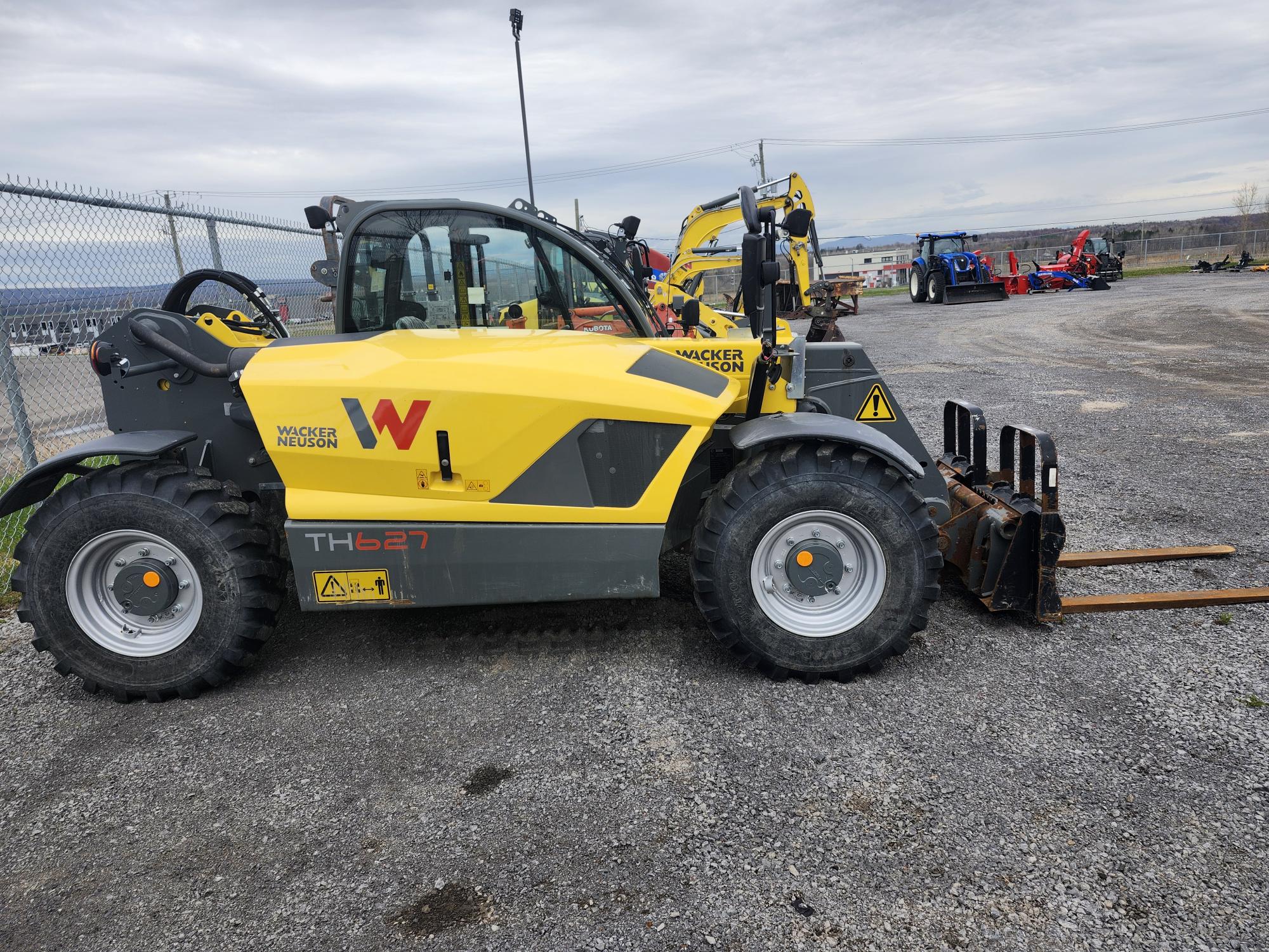 Un chariot télescopique jaune Wacker Neuson TH627 avec de gros pneus est garé sur du gravier près d'une clôture grillagée, avec d'autres équipements de construction visibles en arrière-plan.