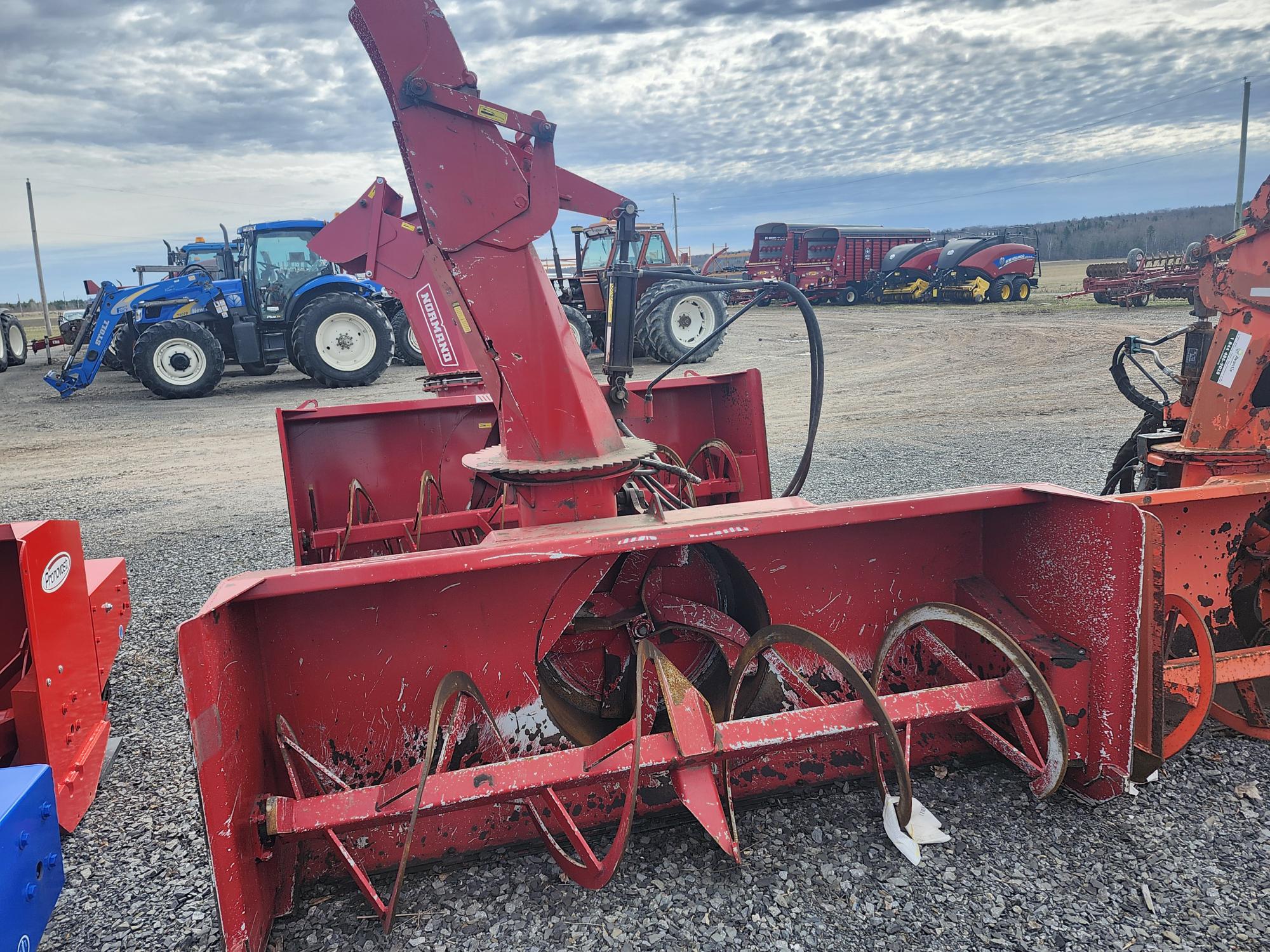 Une grande souffleuse à neige rouge avec des tarières métalliques repose sur du gravier, avec des tracteurs et d'autres équipements agricoles visibles en arrière-plan sous un ciel nuageux.
