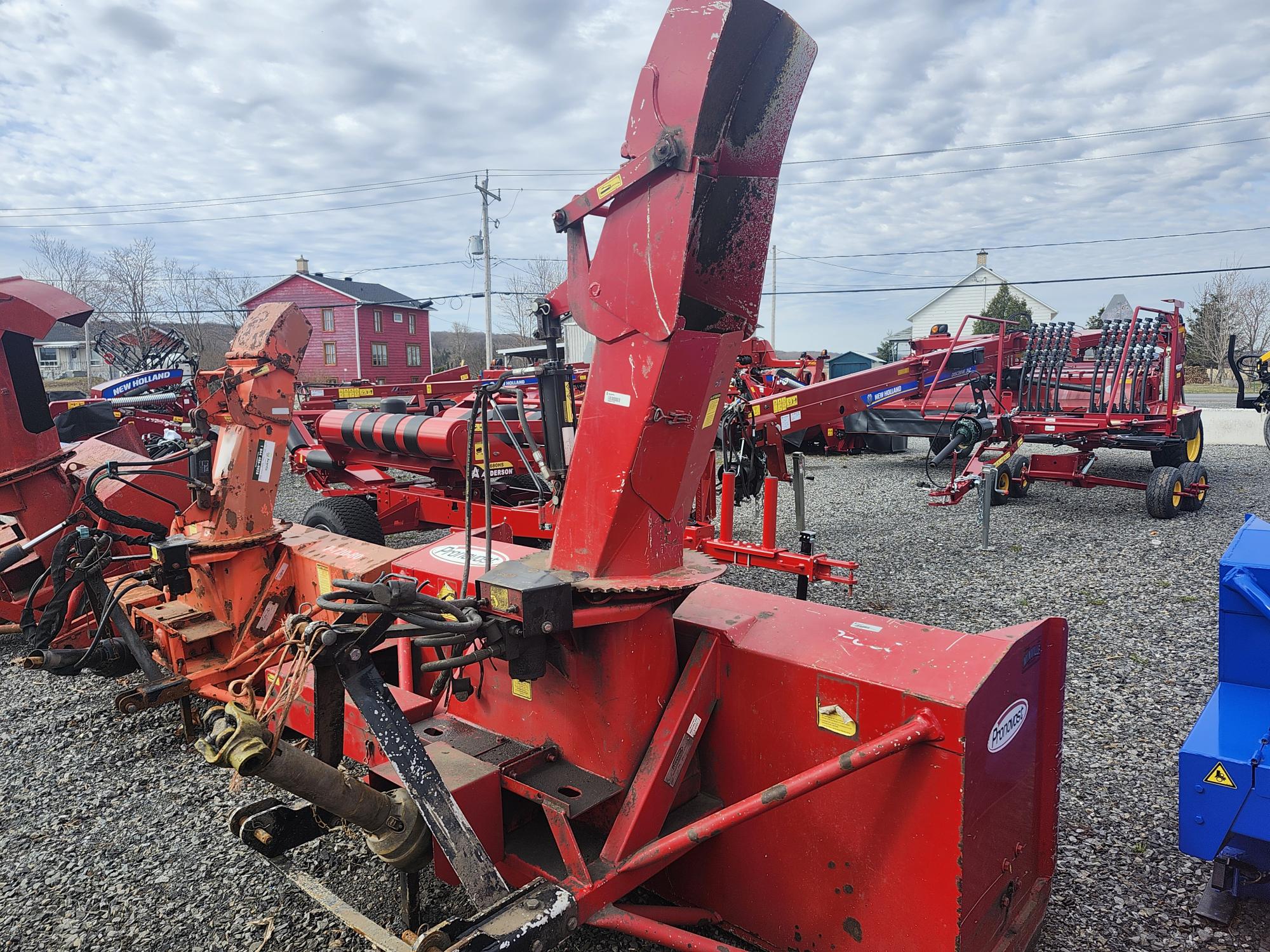 Plusieurs grandes pièces d'équipement agricole rouges sont exposées à l'extérieur sur un terrain en gravier sous un ciel nuageux, avec des granges et des poteaux électriques visibles en arrière-plan.