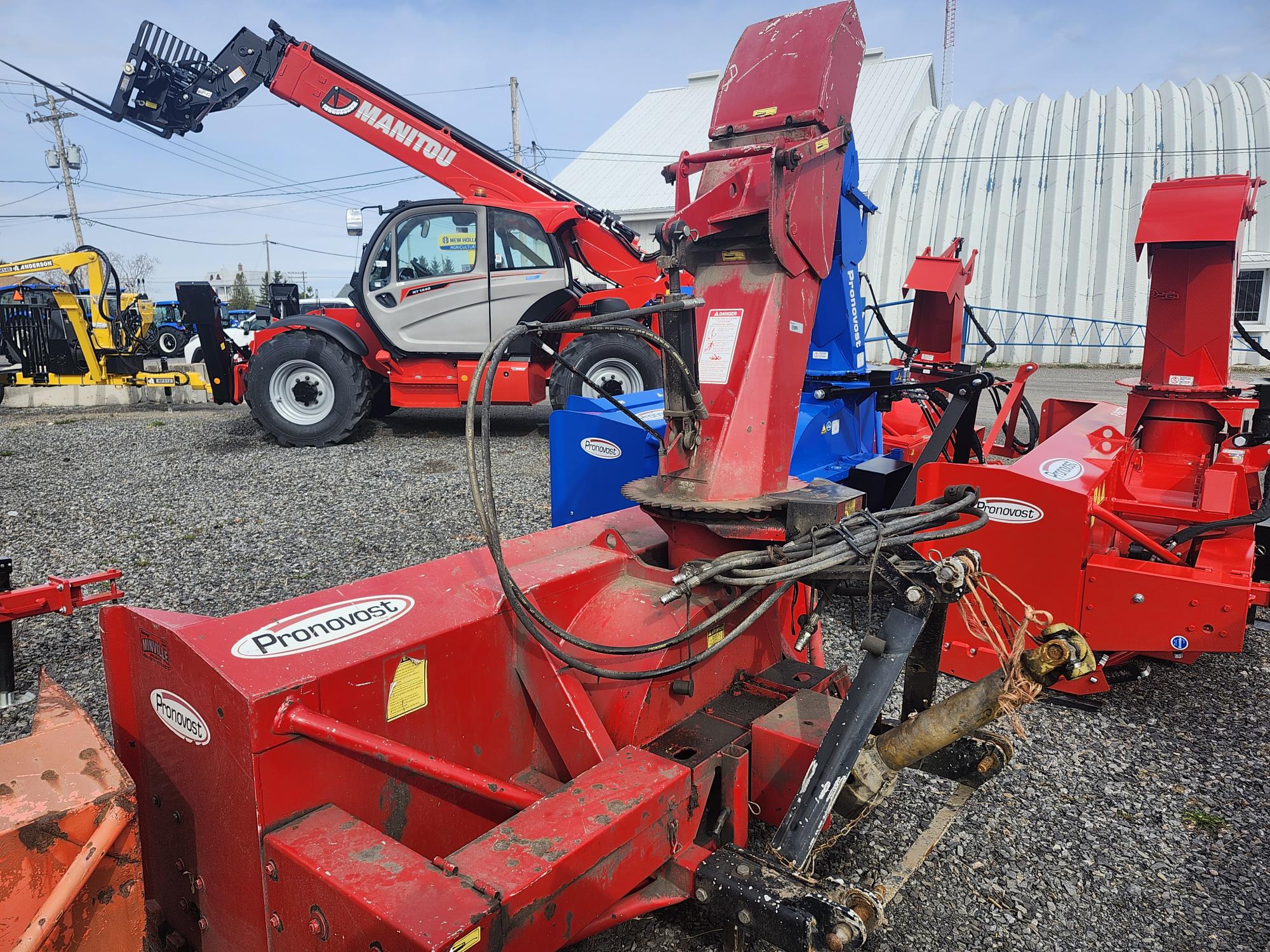 Une souffleuse à neige Pronovost rouge repose sur du gravier, laissant apparaître ses flexibles hydrauliques et ses composants métalliques. À l'arrière-plan, on distingue un chariot télescopique Manitou rouge et d'autres équipements agricoles. Des bâtiments industriels blancs sont également visibles.