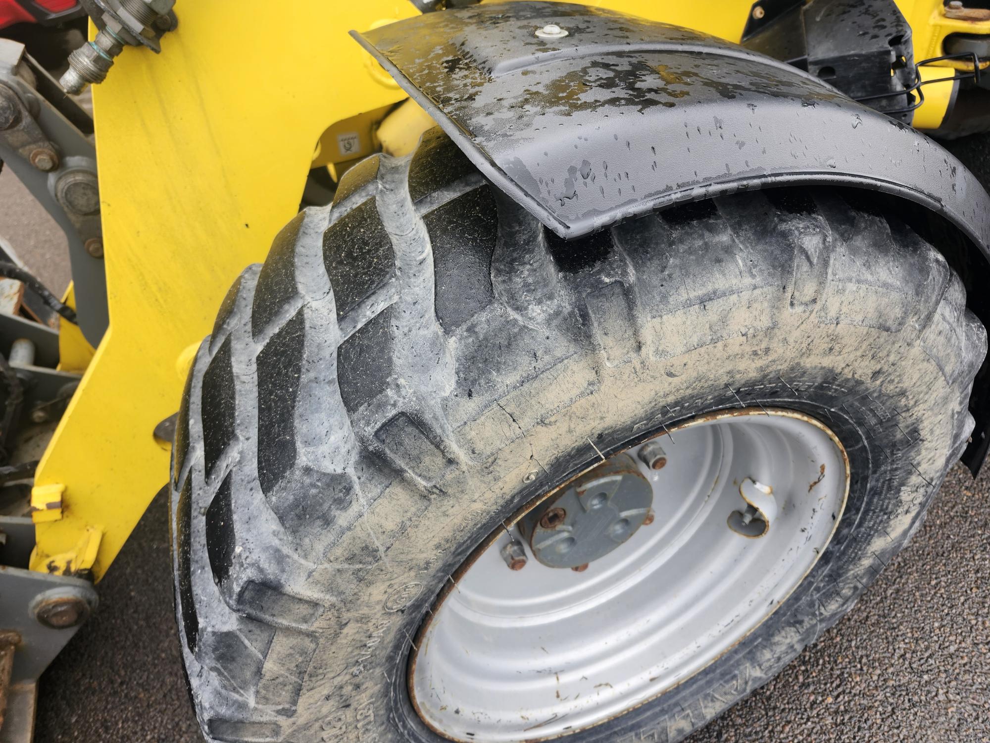 Gros plan d'un gros pneu de tracteur boueux aux sculptures profondes, fixé à un véhicule agricole jaune. Le garde-boue noir au-dessus du pneu est couvert de gouttes d'eau, et le sol semble mouillé.